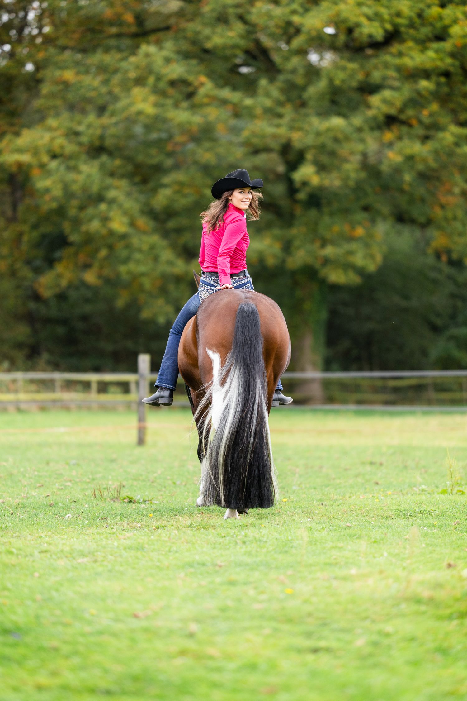 View of horse's tail and back while standing in green field with trees.