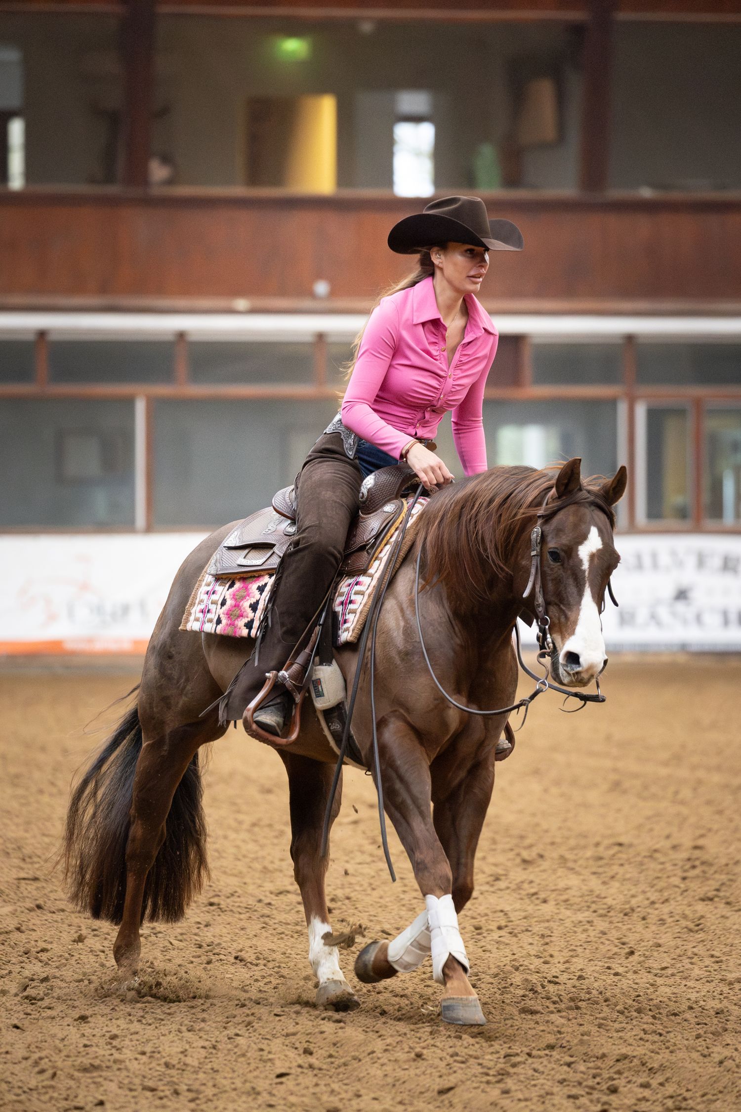 A horse and rider in pink perform a sliding stop maneuver in a sandy indoor arena during a reining competition.