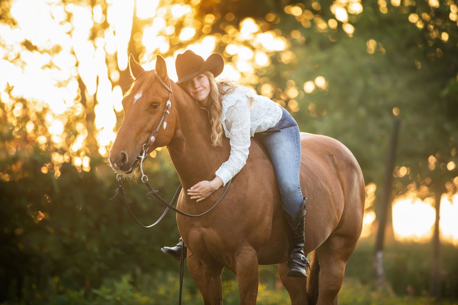 A rider sits atop a brown horse in a sun-dappled field during sunset.