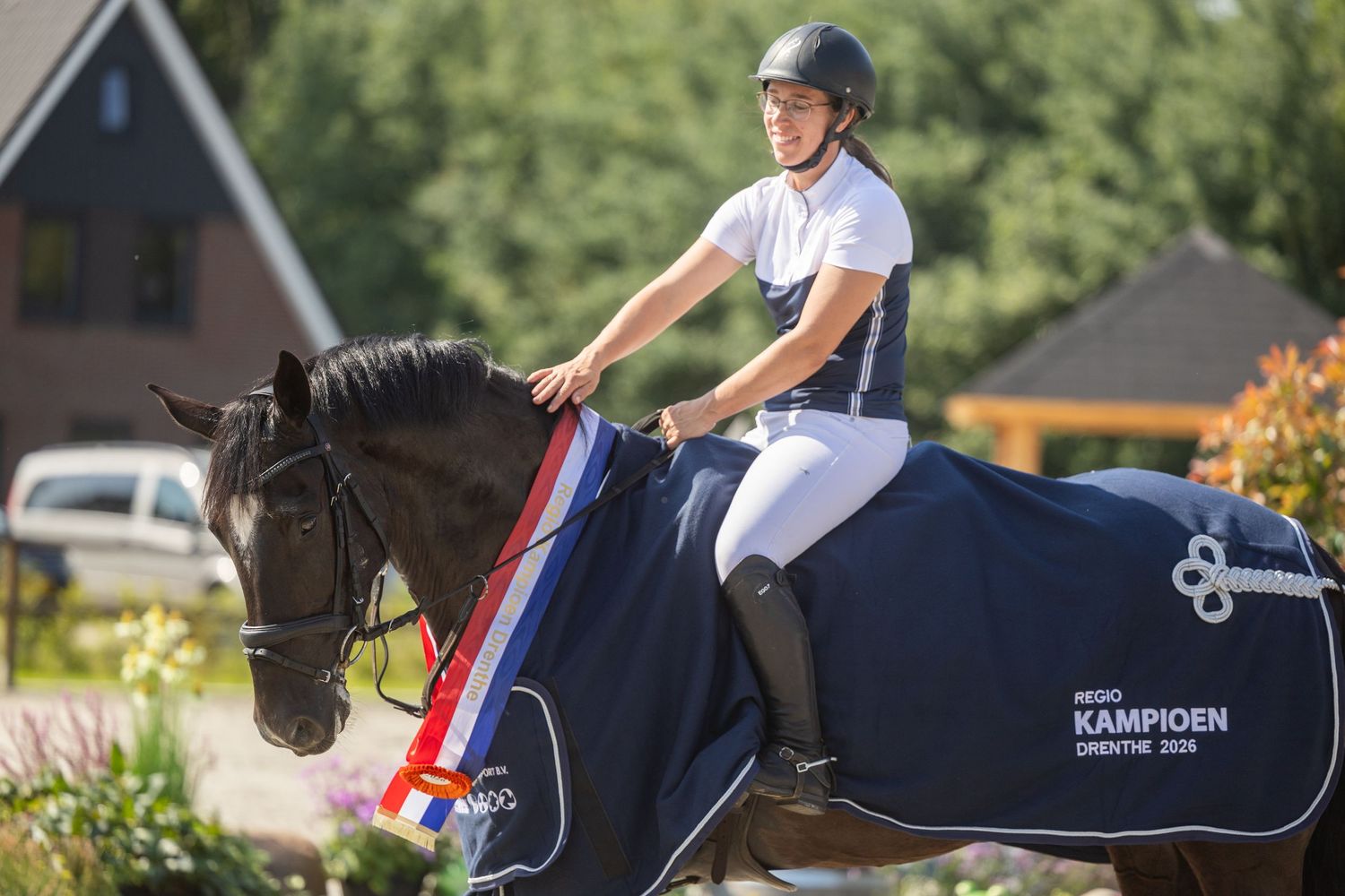 Equestrian rider in white outfit on black horse with champion sash and navy competition blanket at outdoor show.