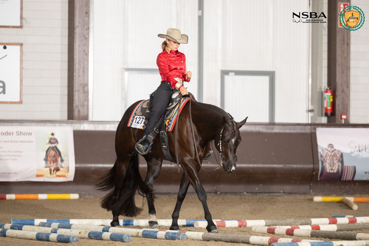 Rider in red jacket on black horse at NSBA equestrian show competition in indoor arena with colorful poles.