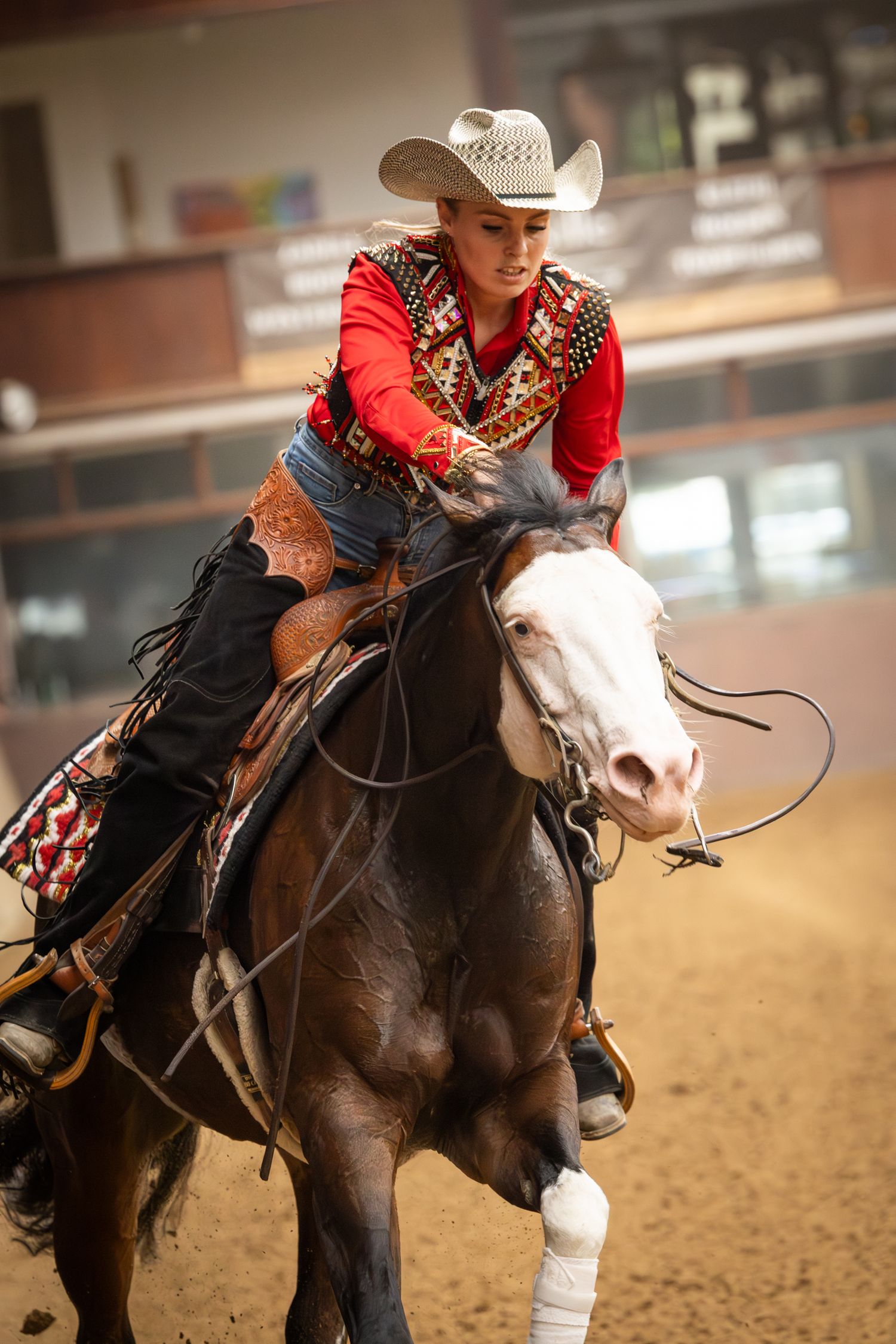Western rider in red decorated shirt competing on a dark horse in an indoor arena.