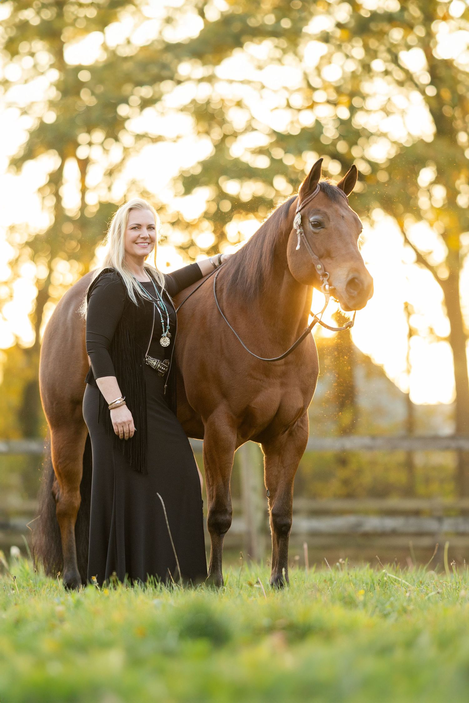 Person in black dress standing with brown horse during golden sunset in field.