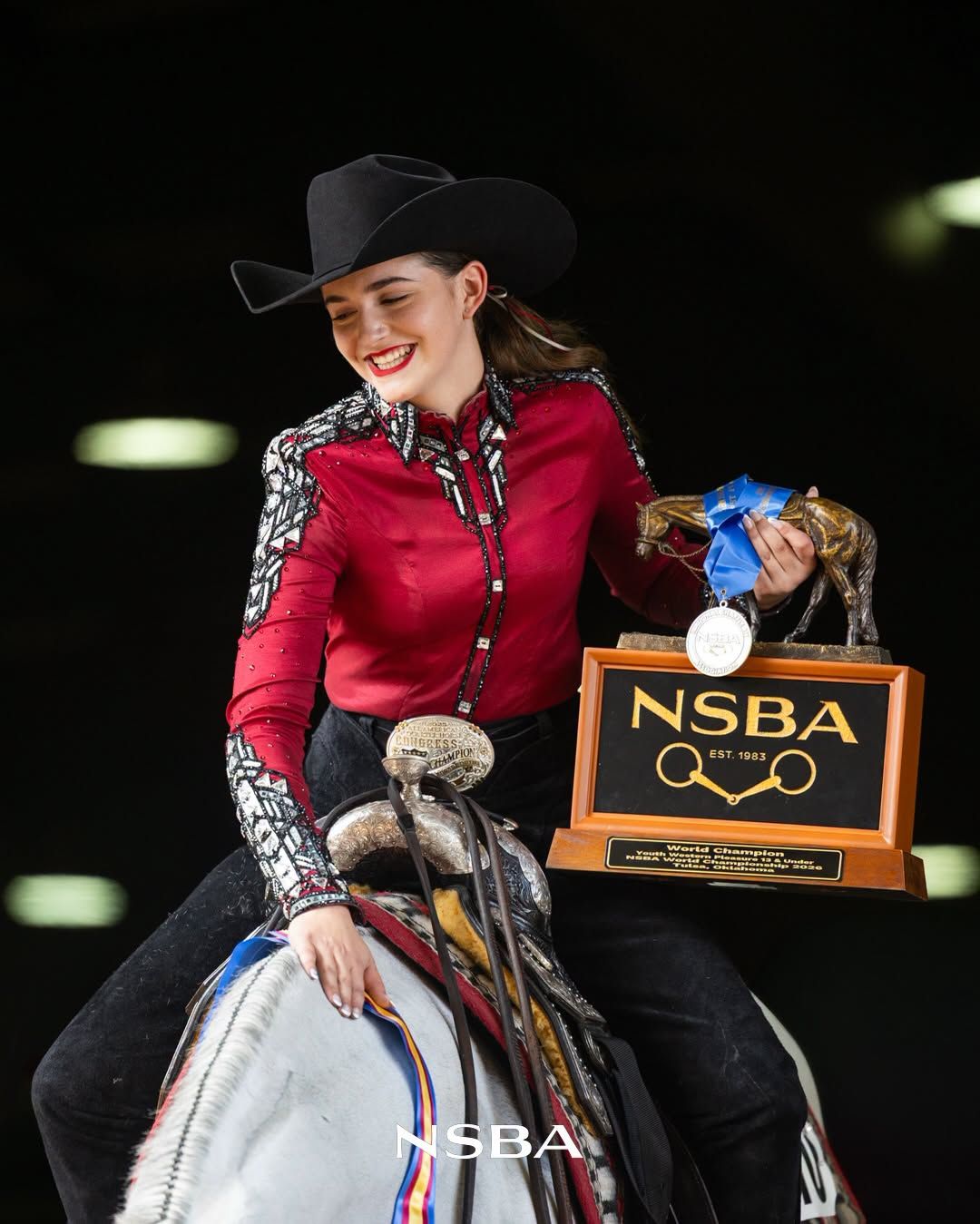 Young woman in red shirt and black cowboy hat holds NSBA trophy while seated on a horse at an equestrian competition.