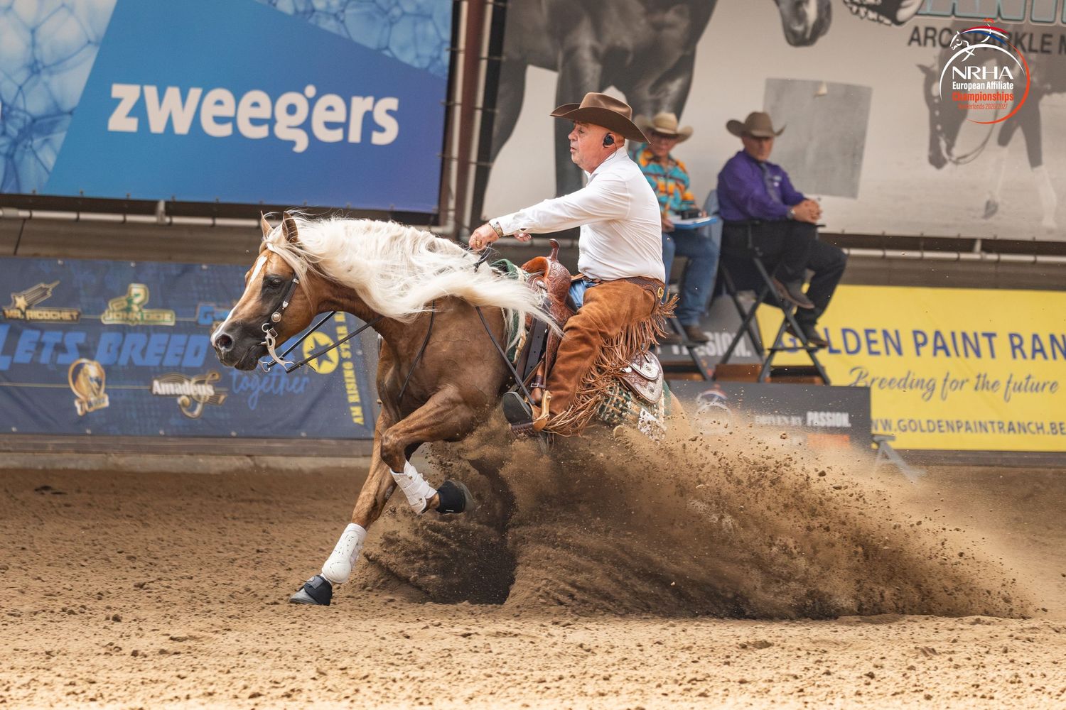A rider on a paint horse performs a sliding stop in a reining competition at an indoor arena with Zweegers signage.