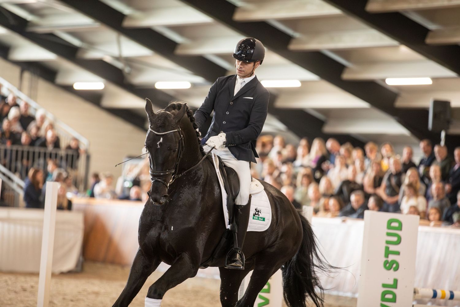 A dressage competition with a rider performing a test on a black horse in an indoor arena with spectators.
