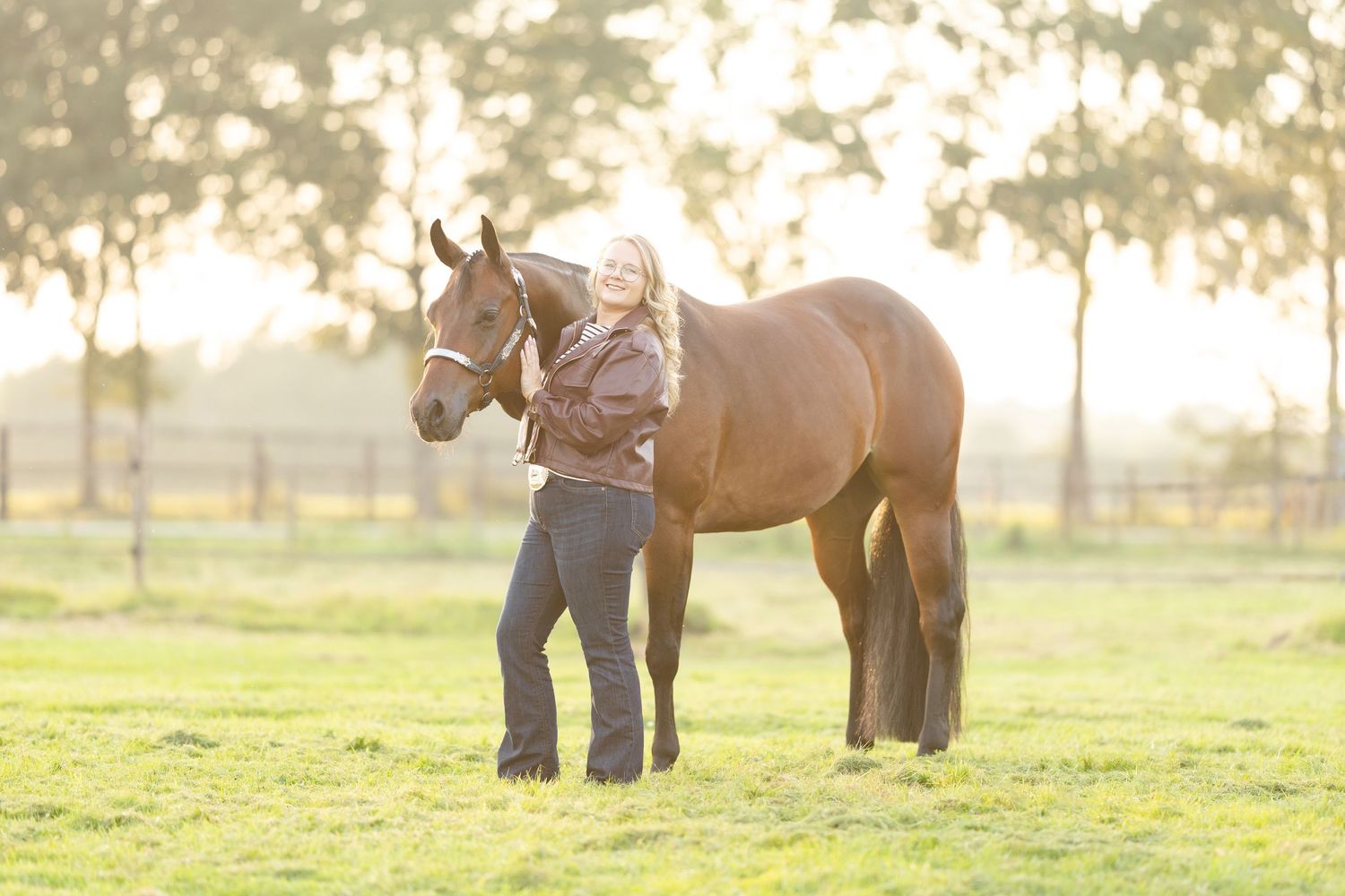 Woman standing with a chestnut horse in a sunlit green pasture with trees in the background.