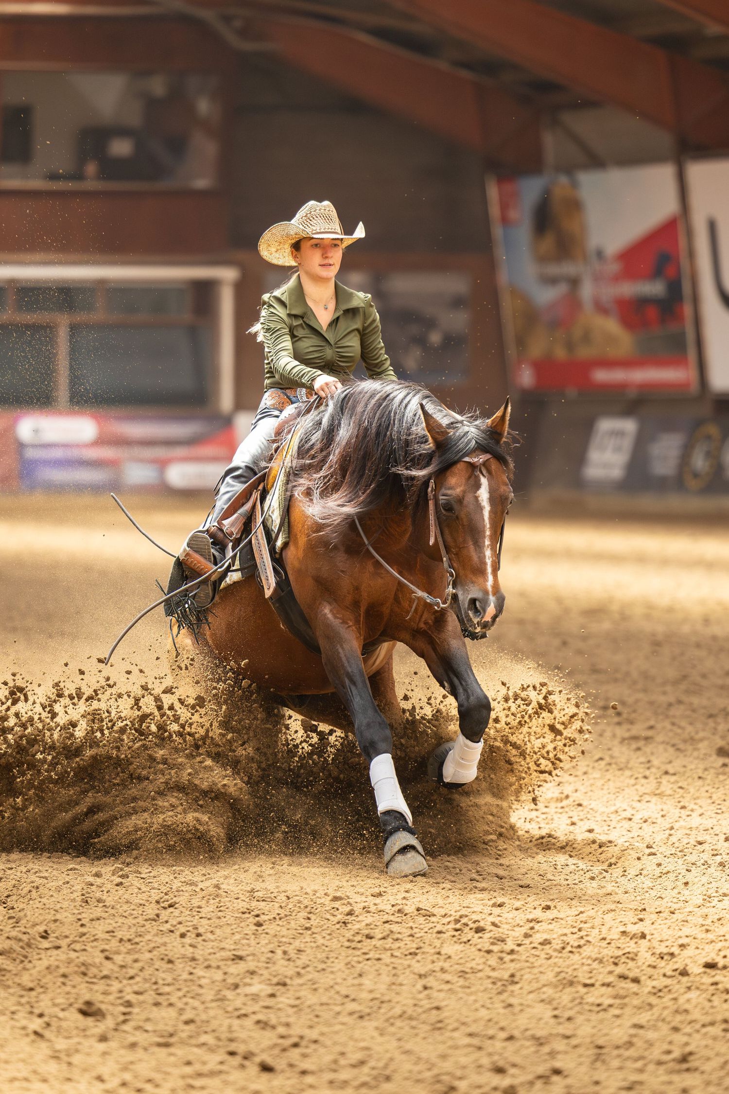 Horse and rider sliding through dirt during reining competition in indoor arena.