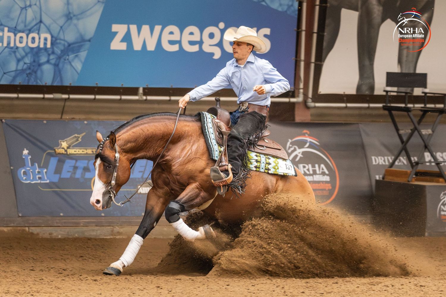 Rider on chestnut horse performing sliding stop in reining competition at Zweegers indoor arena.