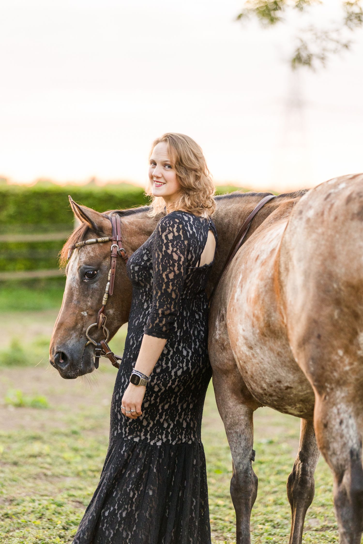 A beautiful dappled horse stands beside someone wearing a black lace dress in a grassy field.