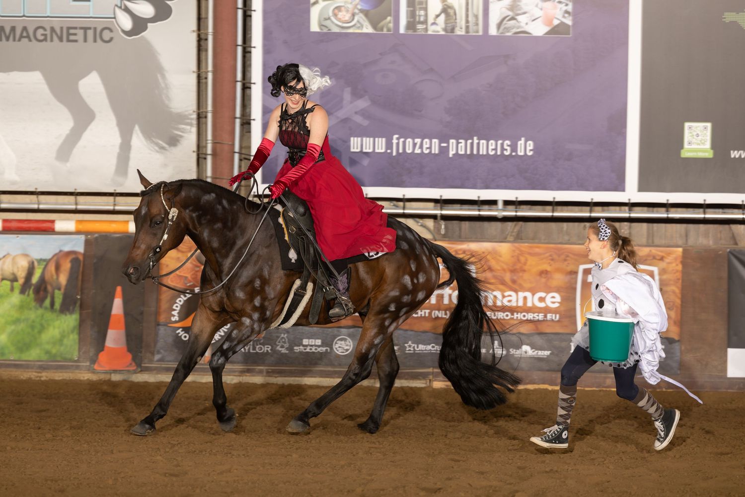 Equestrian in red dress performing side saddle riding while trainer guides the horse in indoor arena.