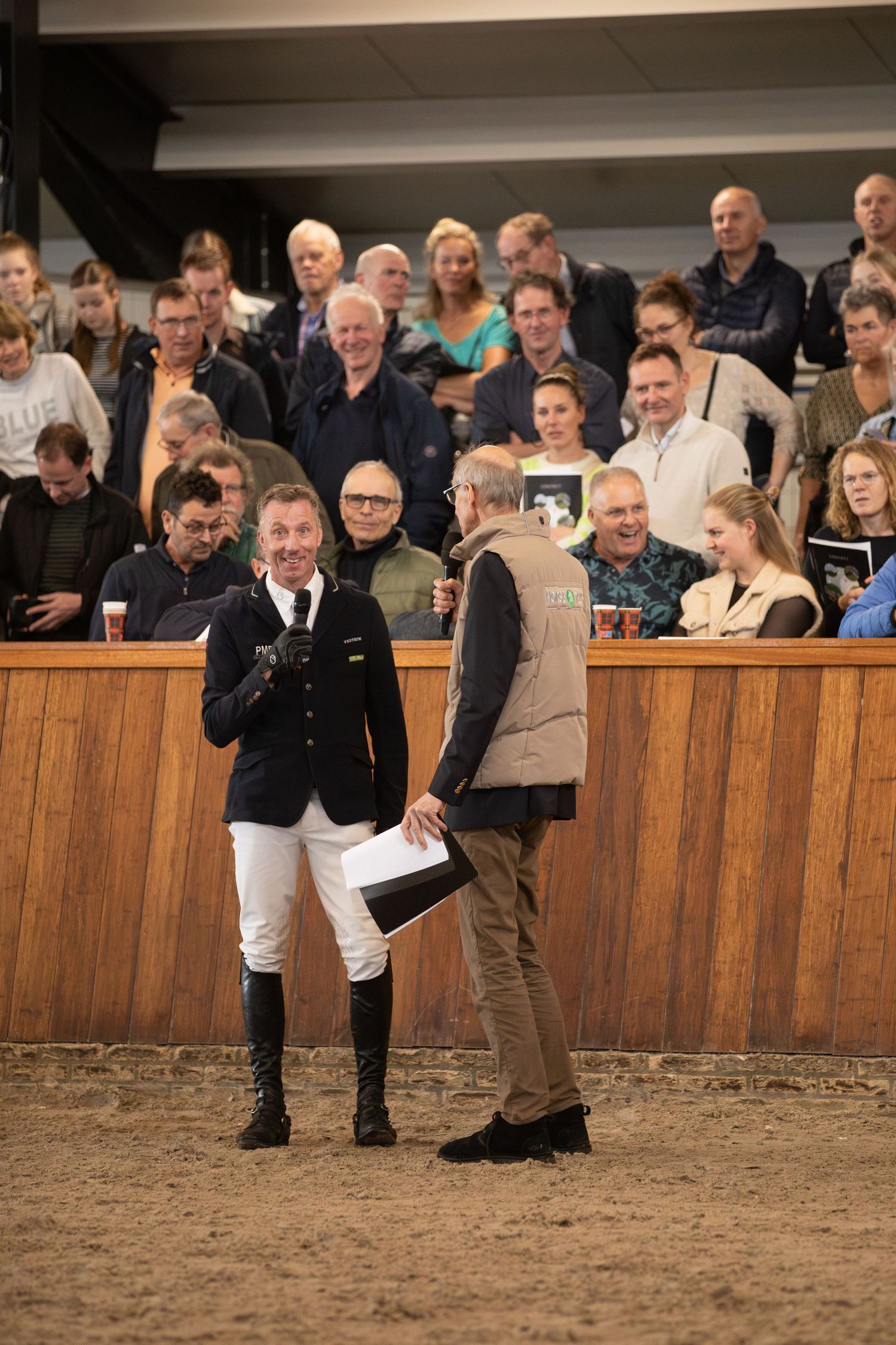 Three people perform in a dirt arena at an equestrian event while spectators watch from behind wooden barriers.