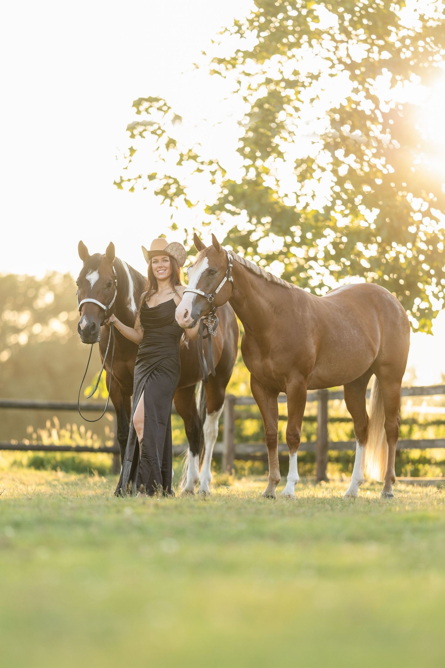 Woman in black dress and cowboy hat stands with two horses at golden hour on a sunny farm.
