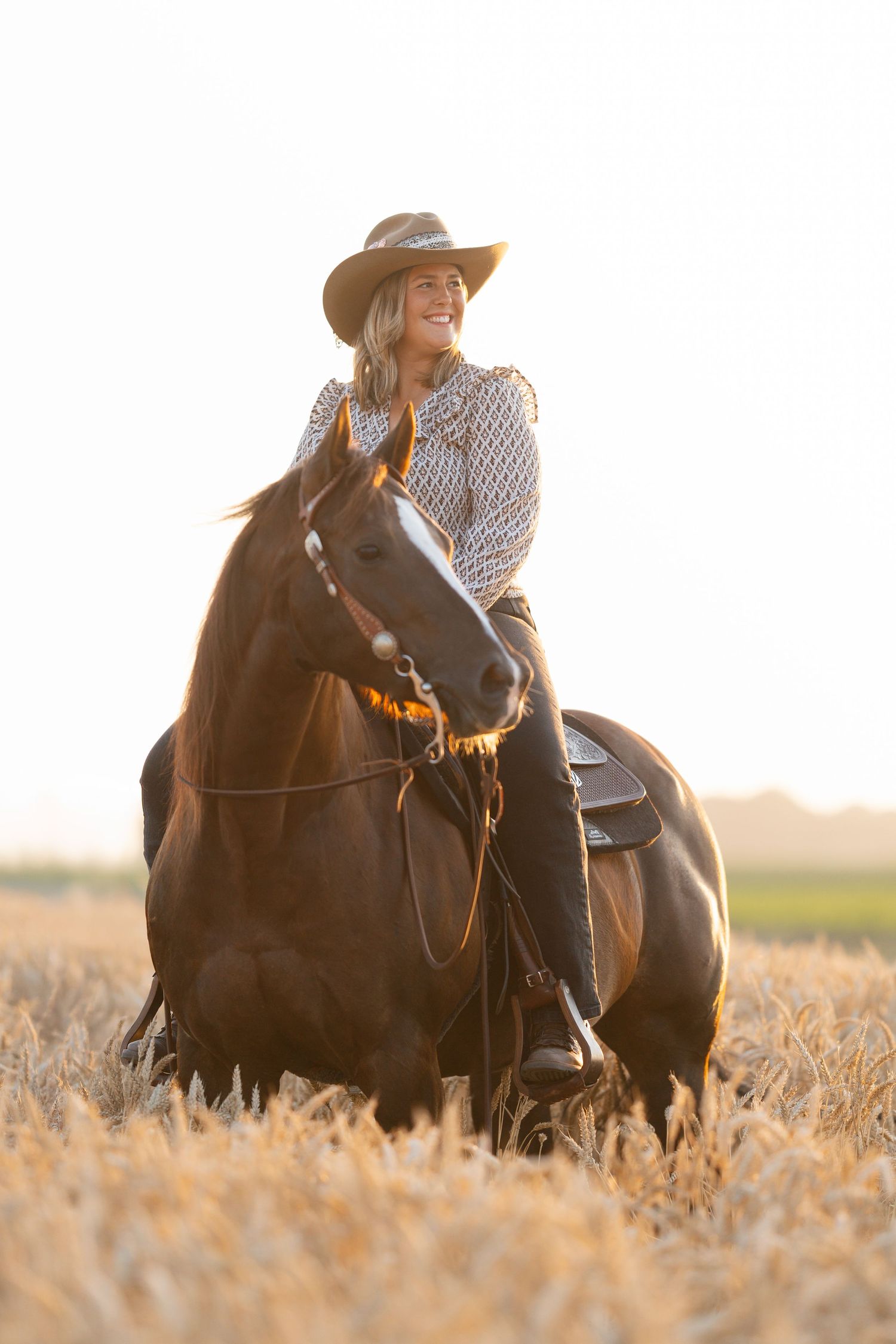 Smiling woman in cowboy hat riding a dark brown horse through a golden wheat field at sunset.
