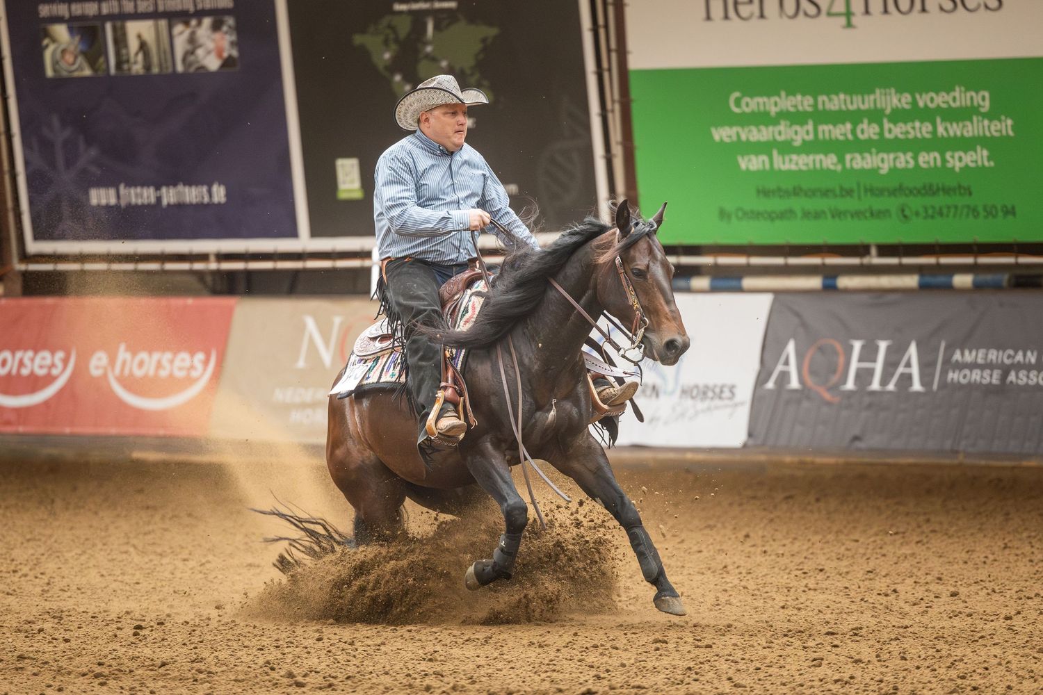 Cowboy riding a dark horse performing a sliding stop in an indoor reining competition on a dirt arena floor.