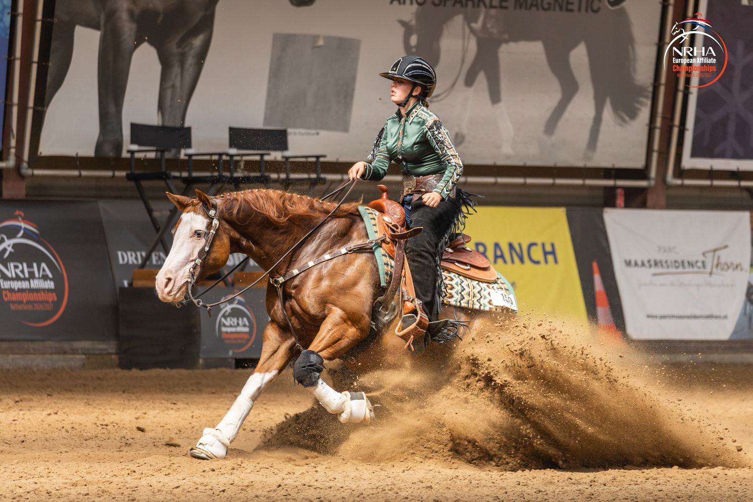Rider on chestnut horse performing sliding stop in NRHA reining competition on indoor dirt arena.