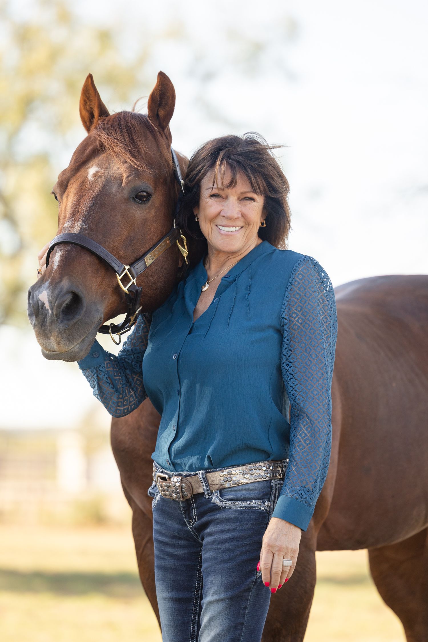 Person in blue top poses cheerfully with brown horse in outdoor setting.