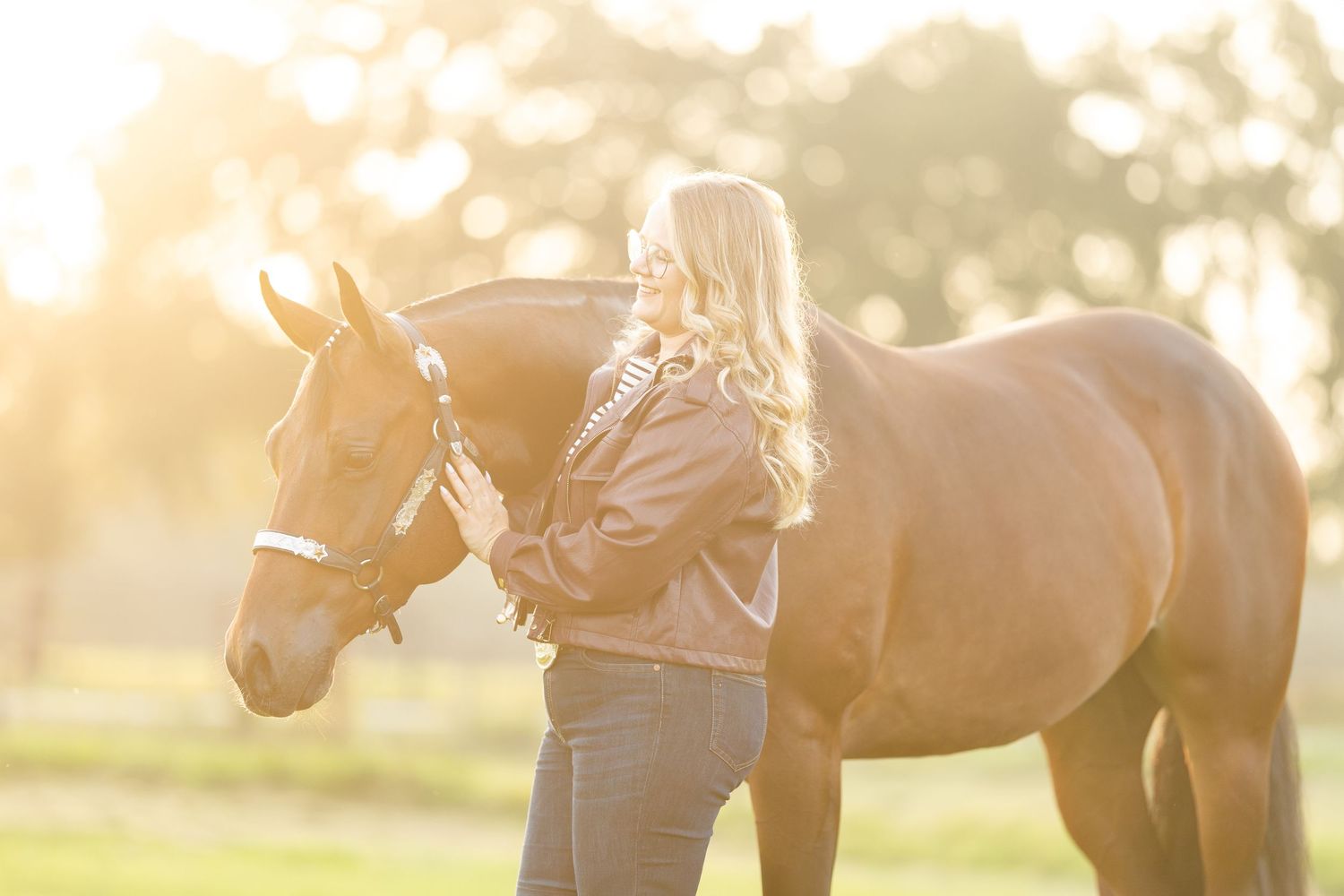 Blonde woman standing beside a chestnut horse in golden sunlight on a green field.