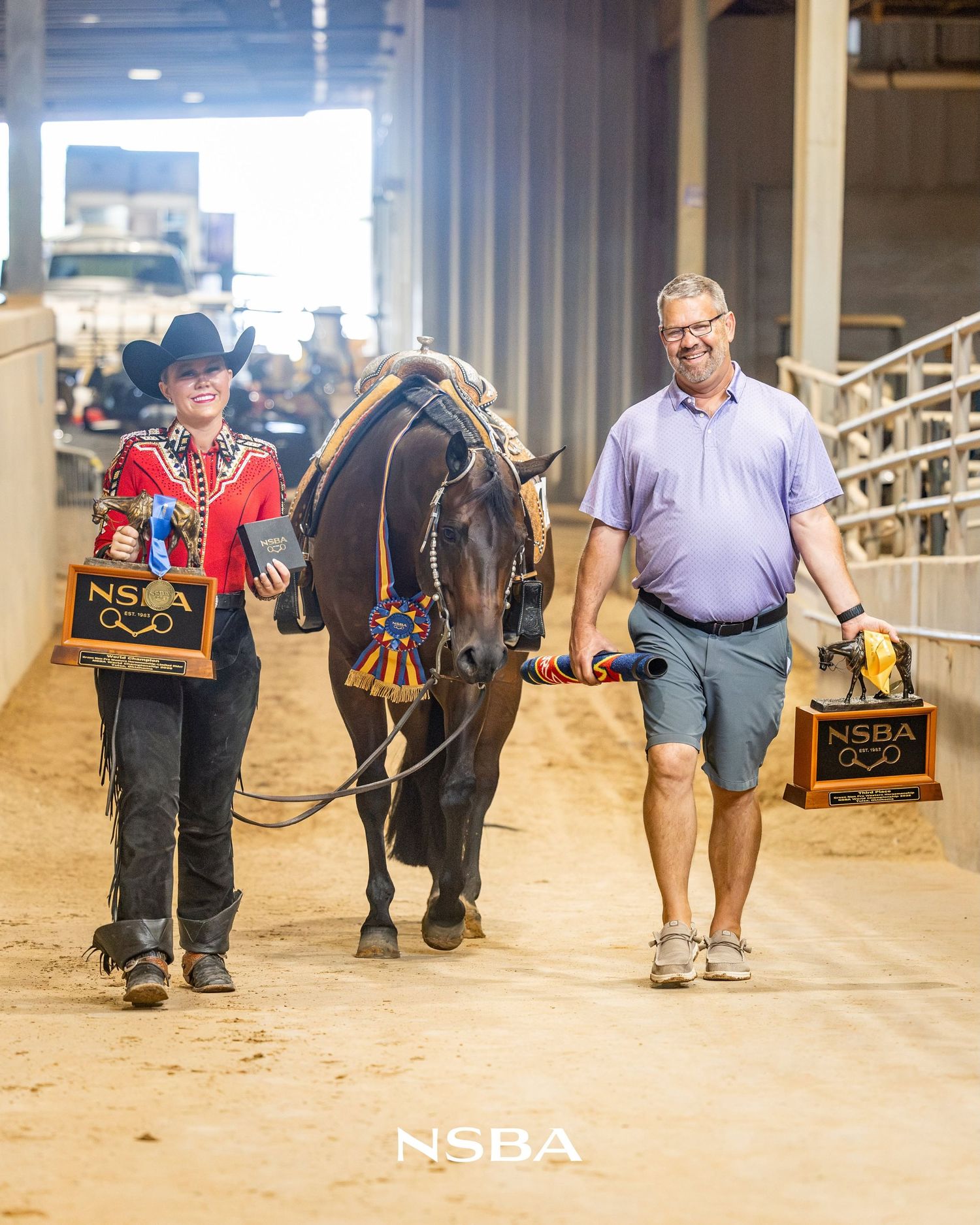 Young rider and man walk horse at NSBA show, holding trophy plaques after winning competition.