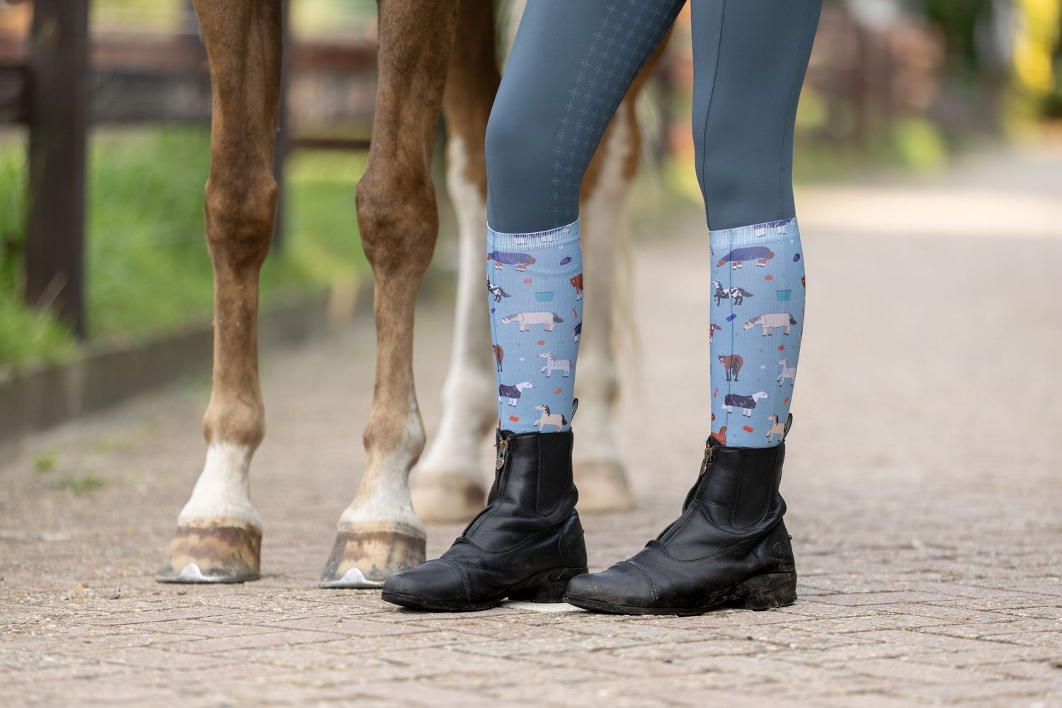 Close-up view of feet and hooves showing equestrian boots next to horse legs on a sandy ground.