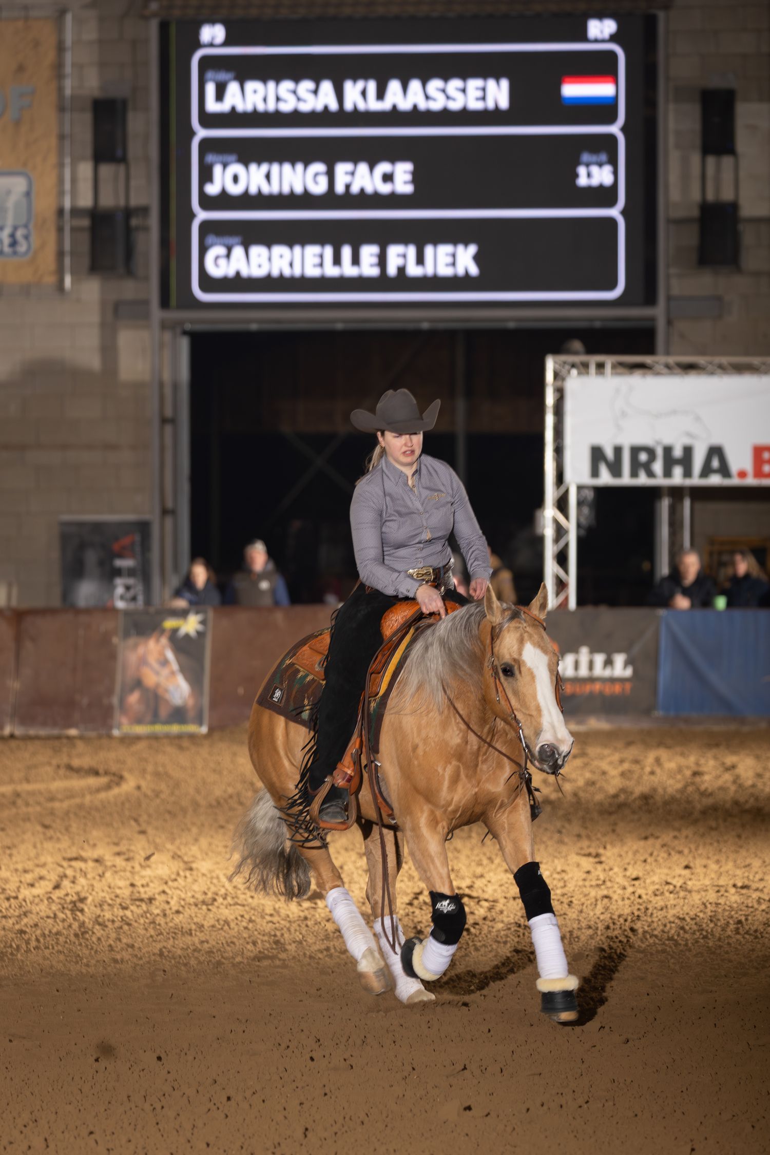 Competitive equestrian riders perform dressage movements in an indoor arena with electronic scoreboard displays.