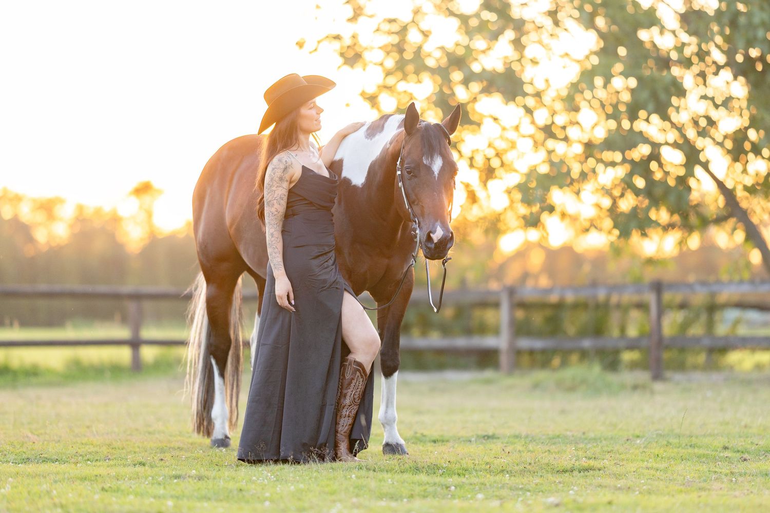 Woman in black gown and hat poses with a paint horse at golden hour in a fenced pasture.