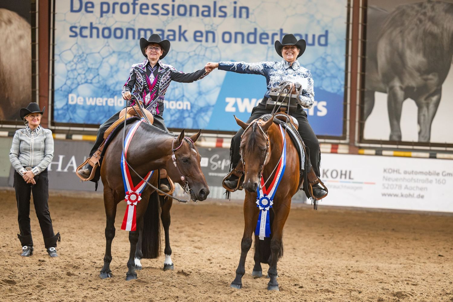 Two riders on decorated horses celebrate wins with ribbons at an indoor equestrian competition in the Netherlands.