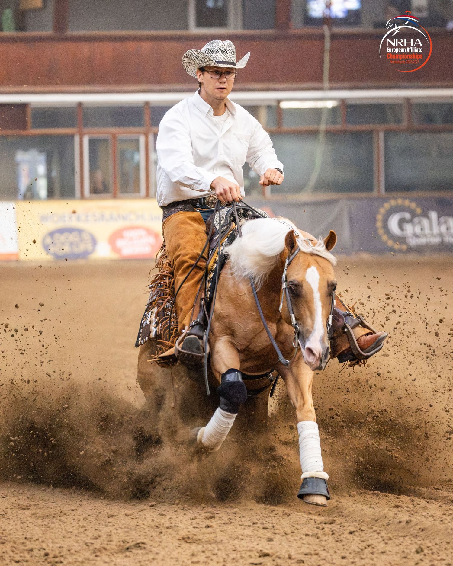 Young cowboy in white shirt and hat performs a sliding stop on a paint horse in a reining competition.