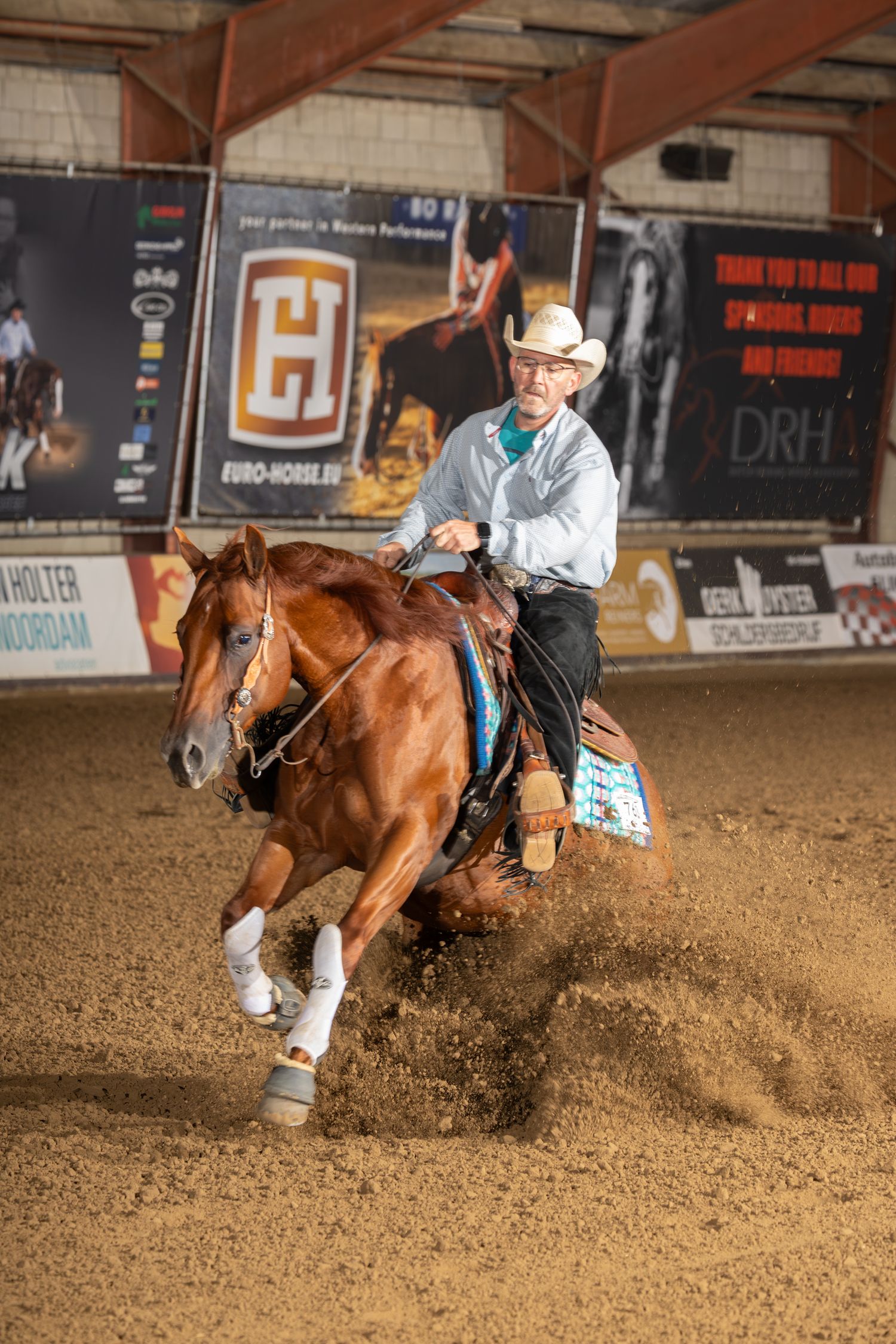 A horse and rider perform a sliding stop maneuver in a sandy arena during a reining competition.