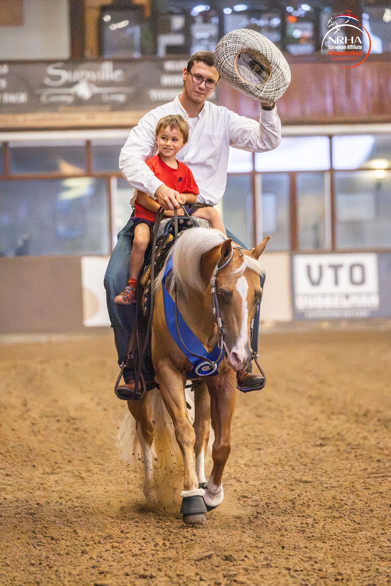 Man and child riding a palomino horse at an NRHA reining competition, man waving cowboy hat in celebration.