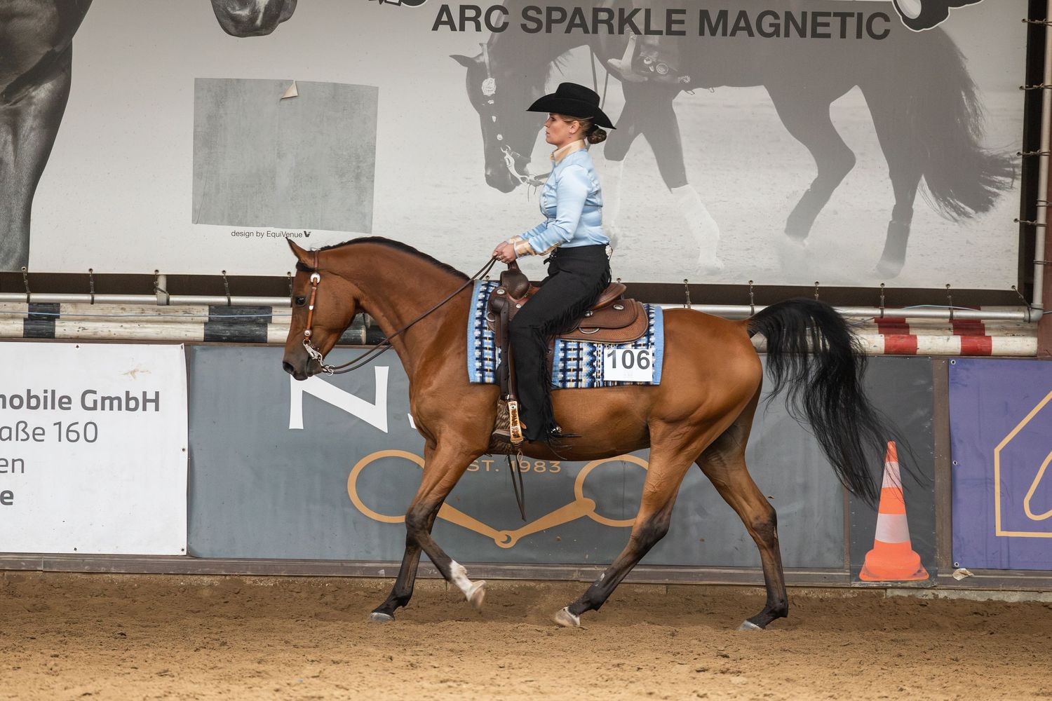 Rider on chestnut horse competing in western equestrian event indoors, wearing blue top, number 106 visible.