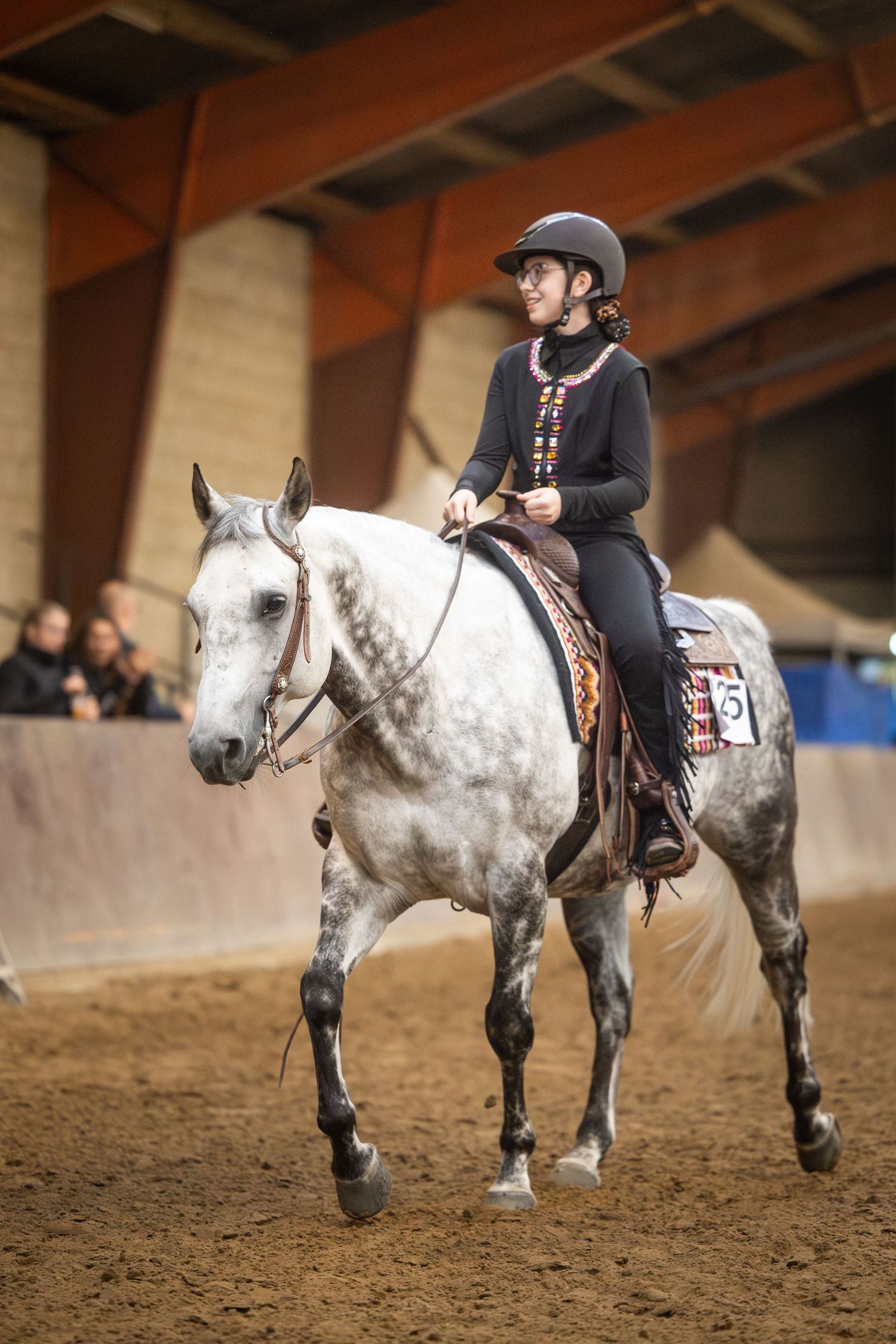 Young woman in black outfit and helmet riding a dapple gray horse in an indoor equestrian arena.