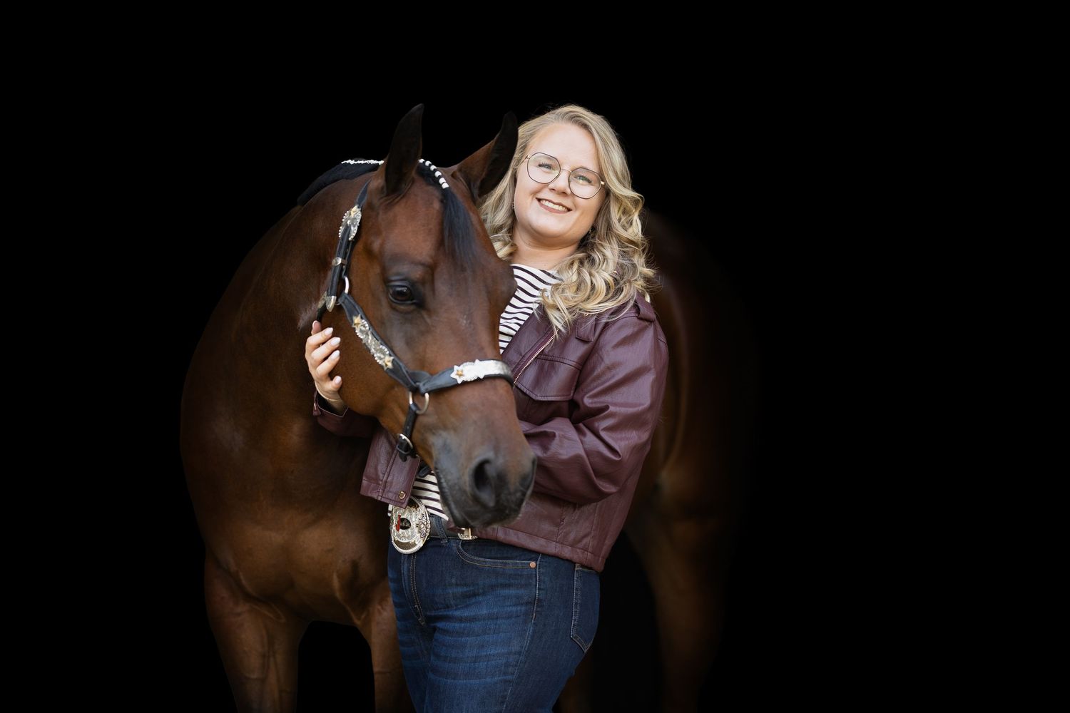 Young blonde woman in burgundy jacket smiling while embracing a brown horse against a dark background.