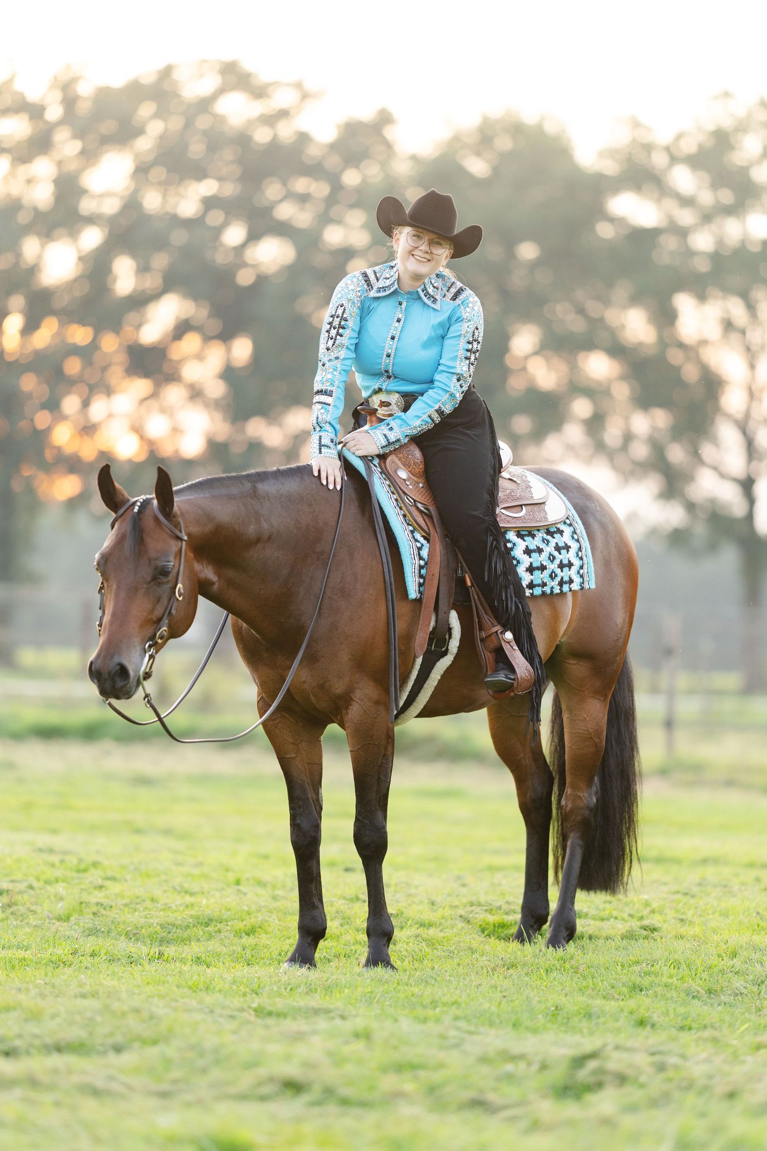 Woman in turquoise western shirt and cowboy hat riding a bay horse on green pasture at golden hour.