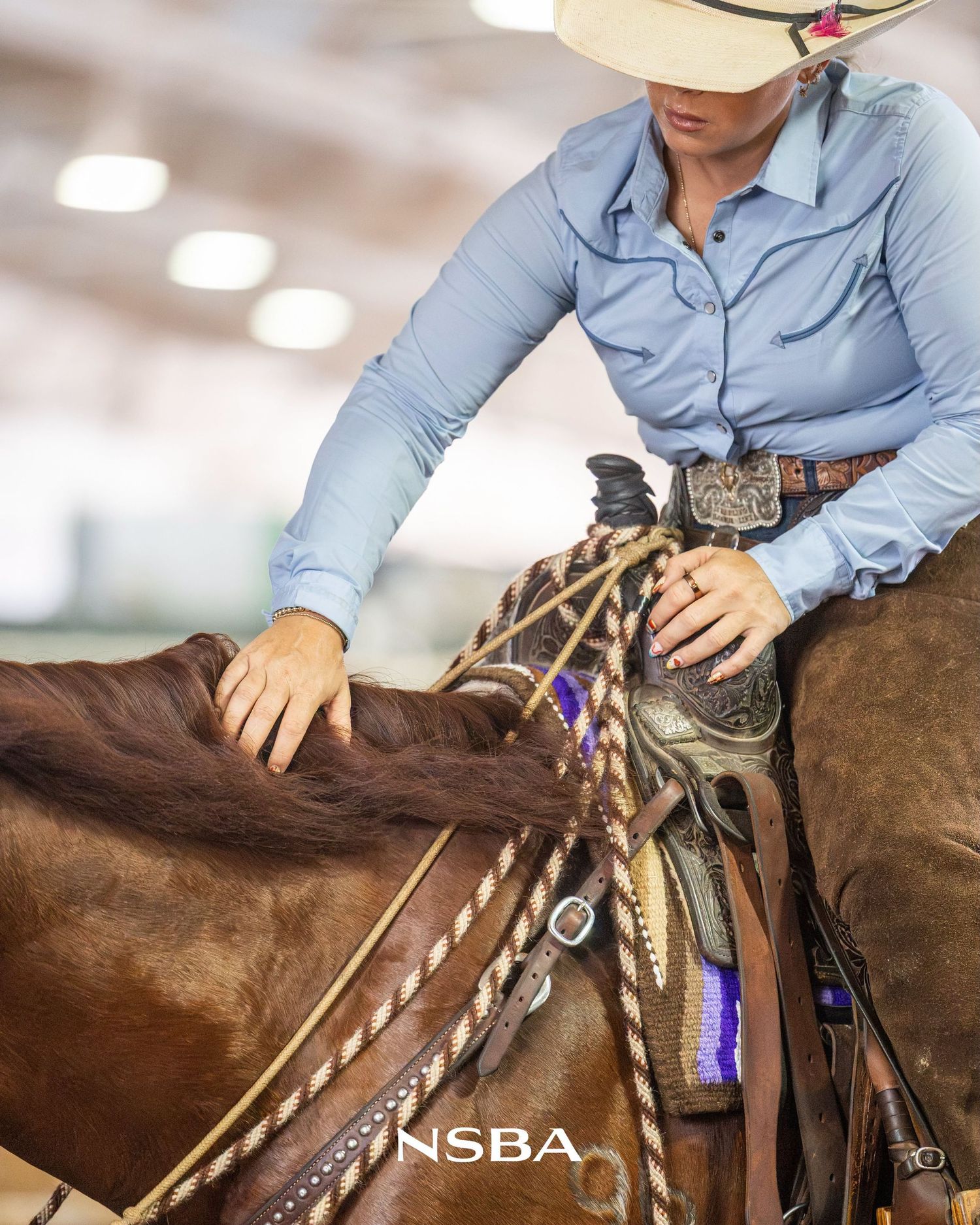 Western rider in blue shirt pats brown horse at NSBA equestrian competition indoors.