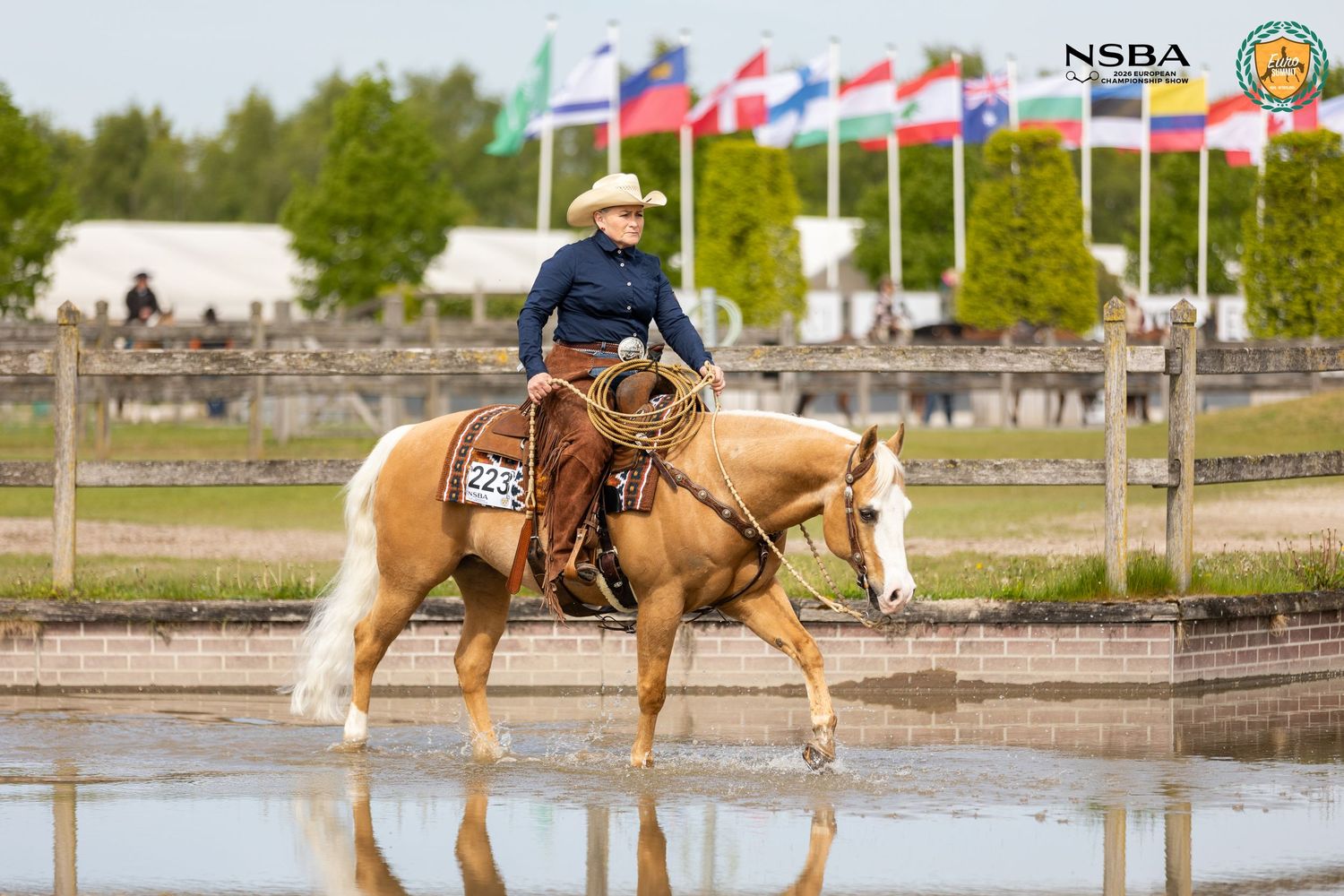 Rider on palomino horse competing at NSBA equestrian event, reflected in water on arena ground.