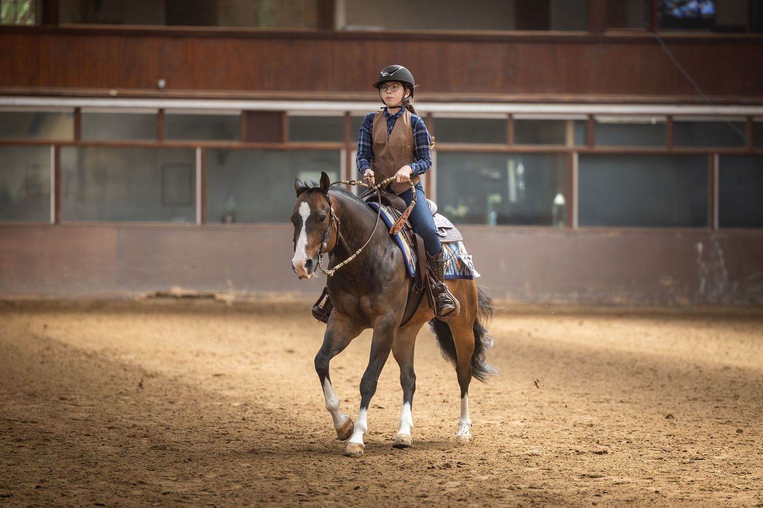 A rider in a helmet guides a brown horse through an indoor equestrian arena with sandy footing.