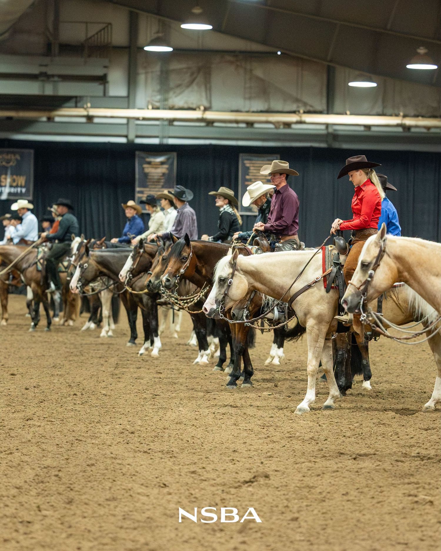 Riders on horses line up in an indoor NSBA equestrian arena during a western horse show competition.
