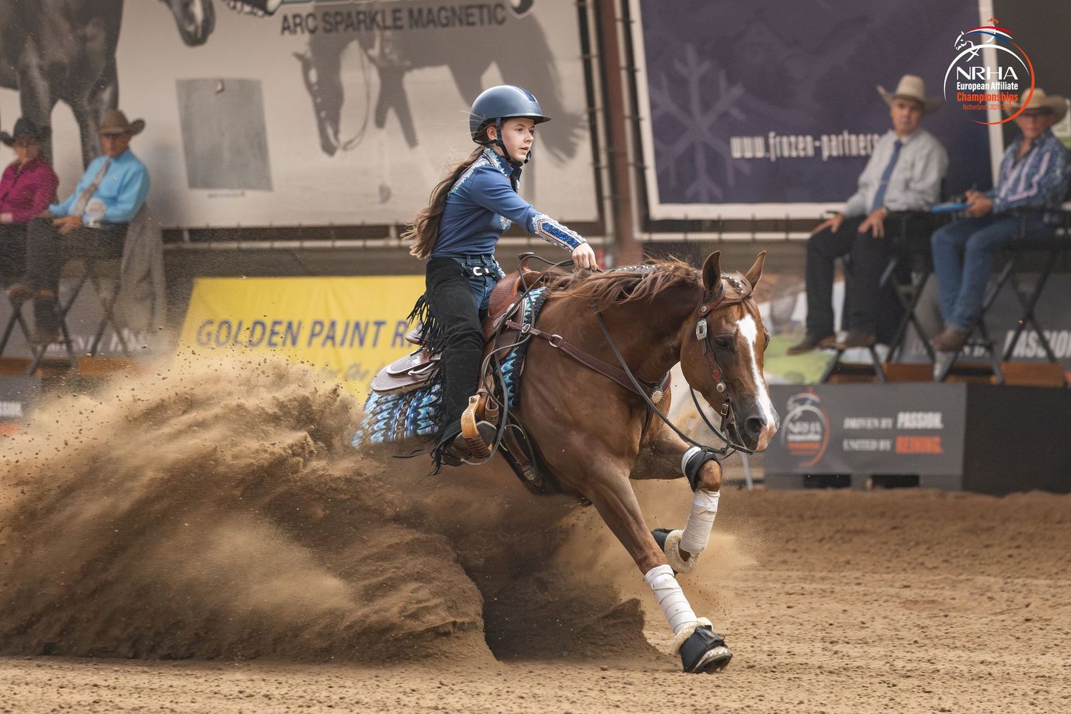 A rider on a chestnut horse performs a sliding stop in a reining competition, kicking up dust in an indoor arena.