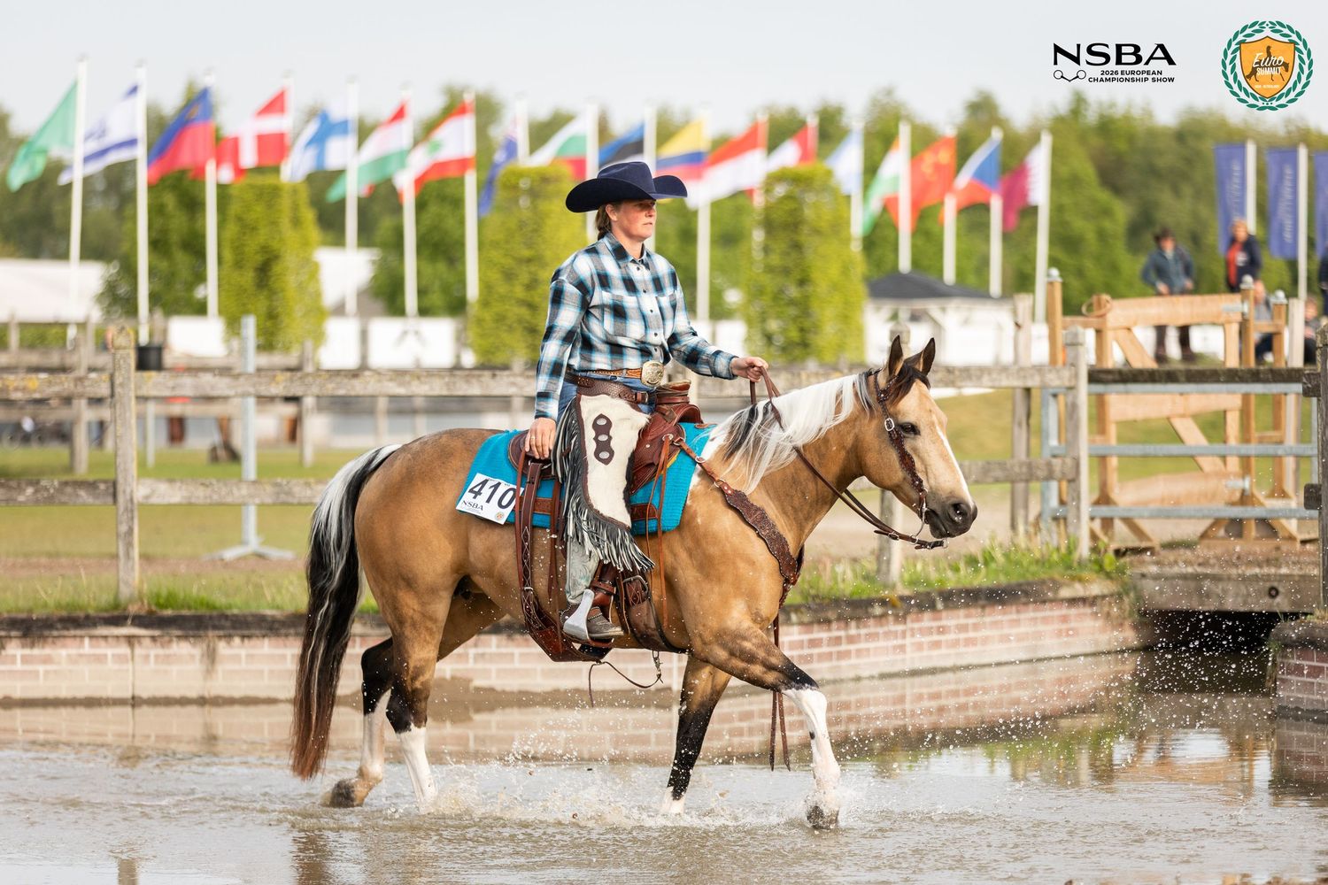 A rider in a plaid shirt competes on a buckskin paint horse at an NSBA outdoor equestrian event.