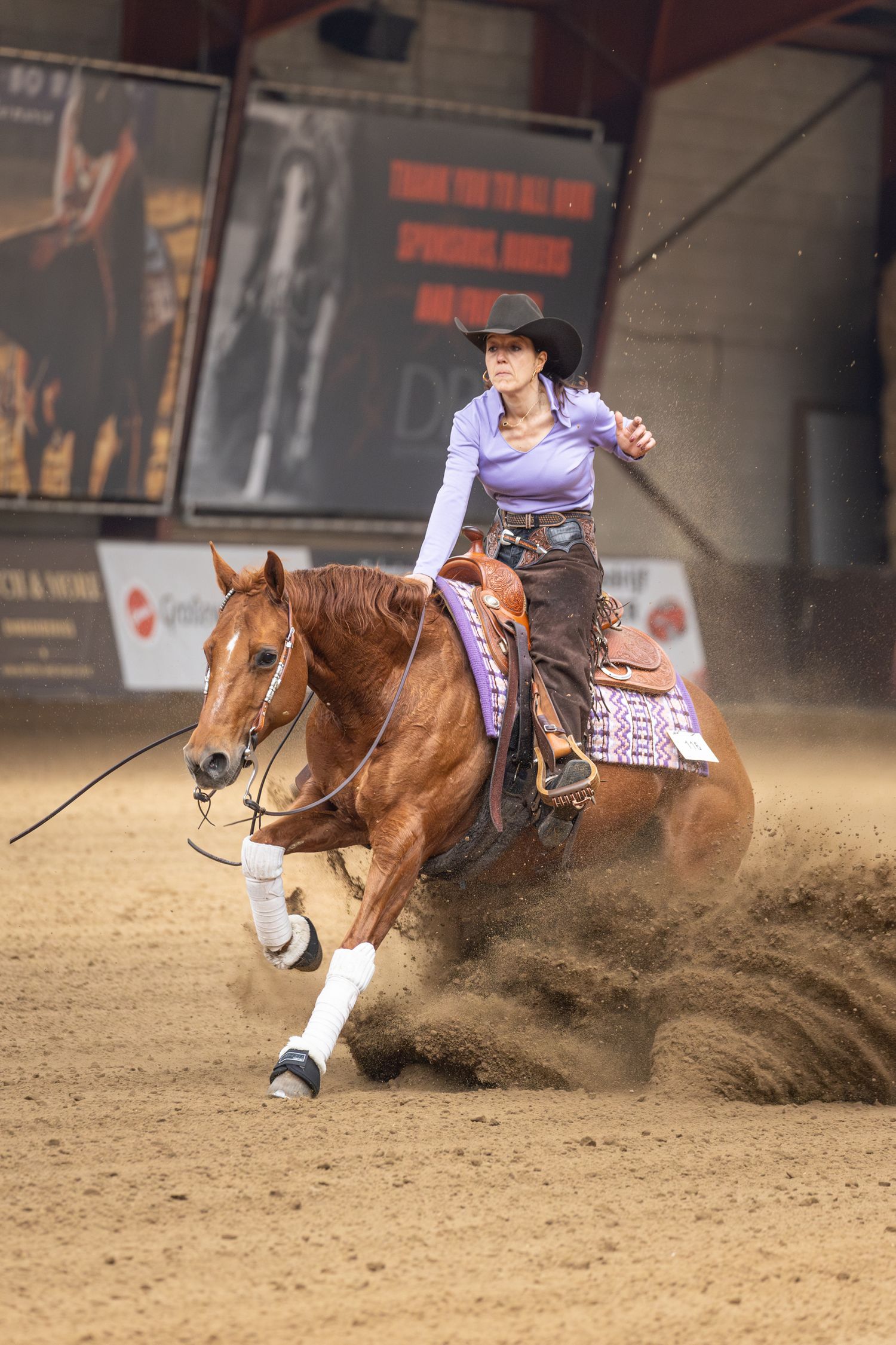 A chestnut horse and rider perform a reining slide maneuver in an indoor arena, kicking up dust.