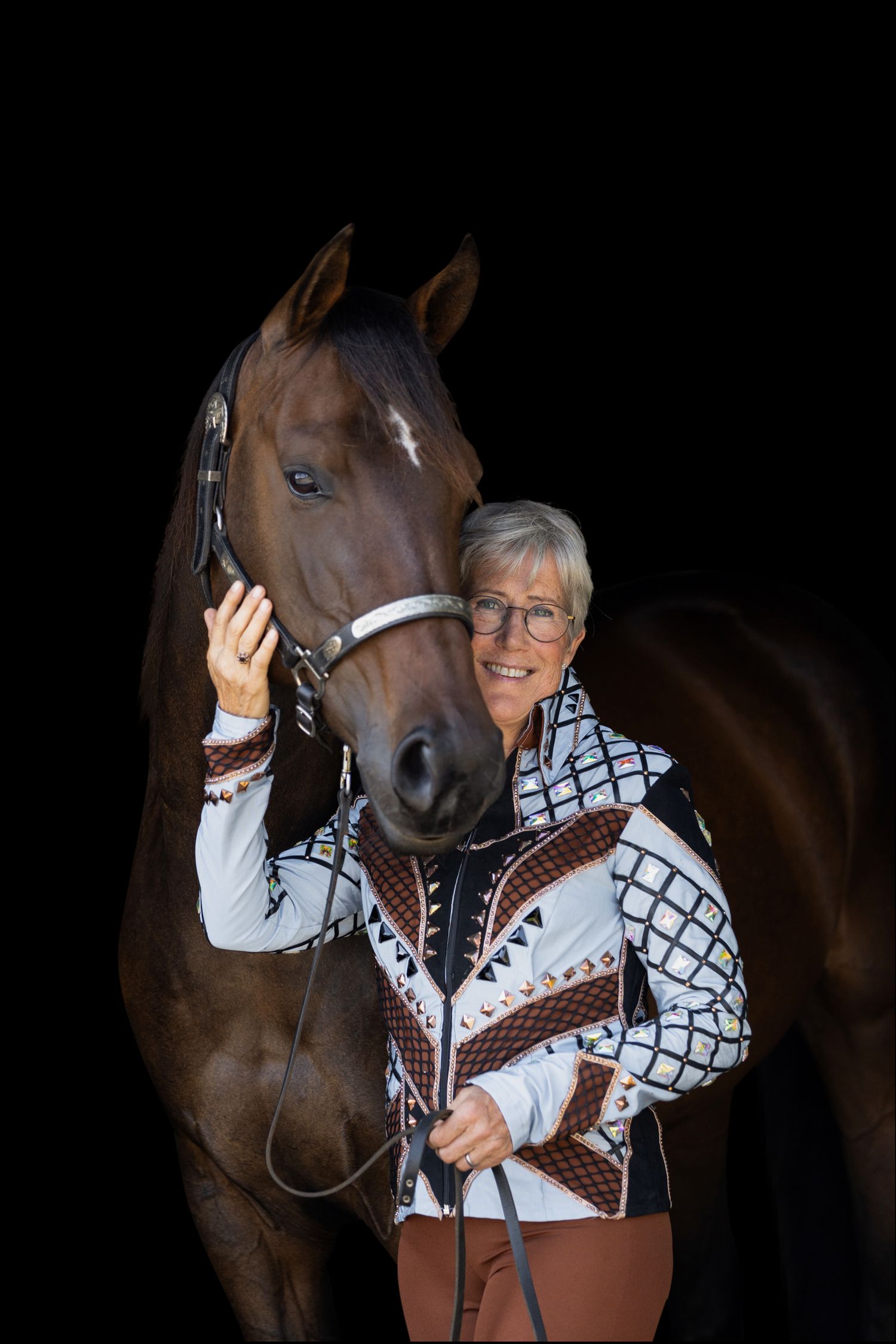 Person in western-style outfit interacting with a brown horse against black background.