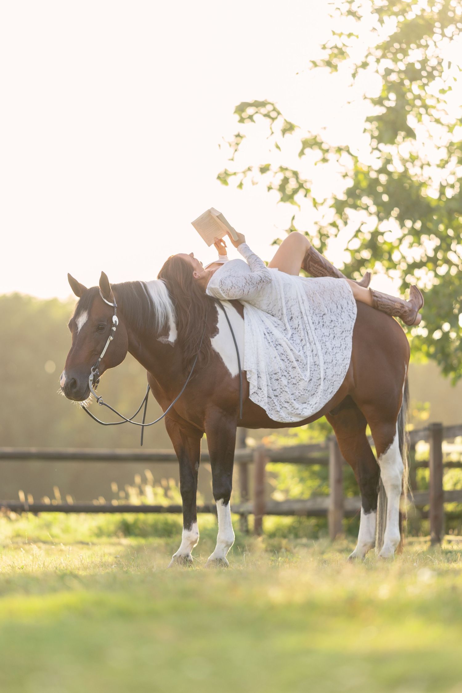 Woman in white lace dress lying on horseback reading a book in a sunlit pastoral field.