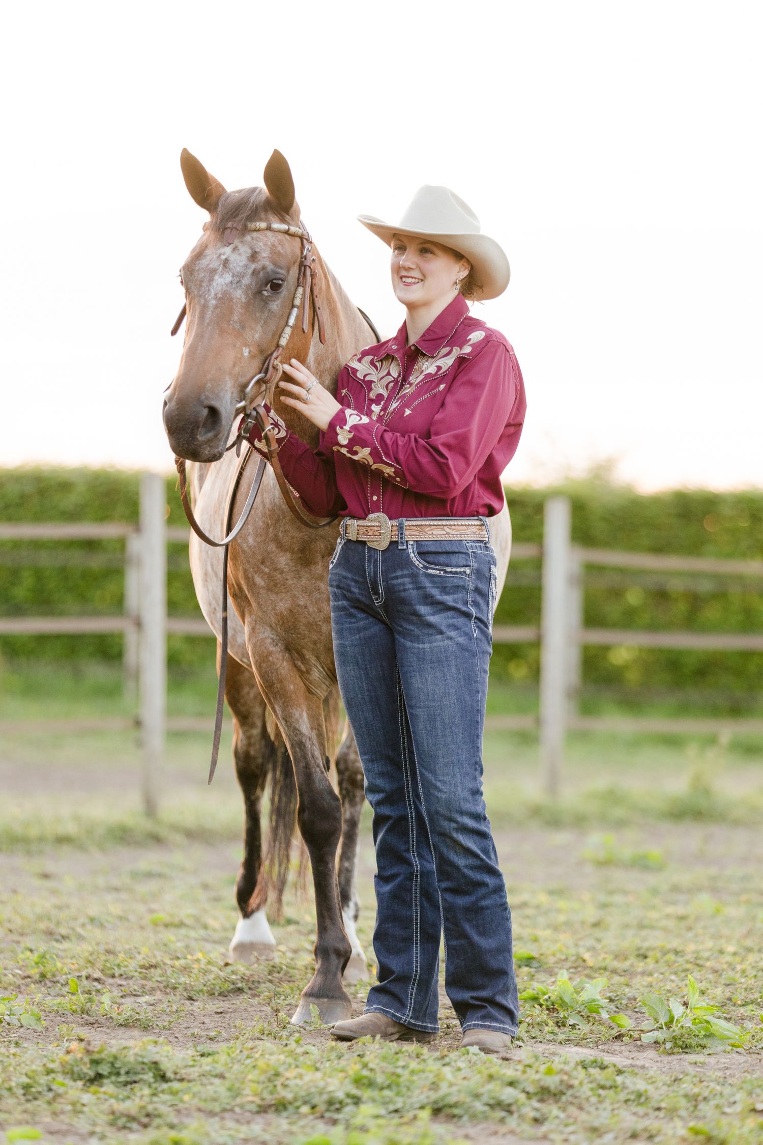 A dappled horse poses with someone in western attire against a fenced field background.