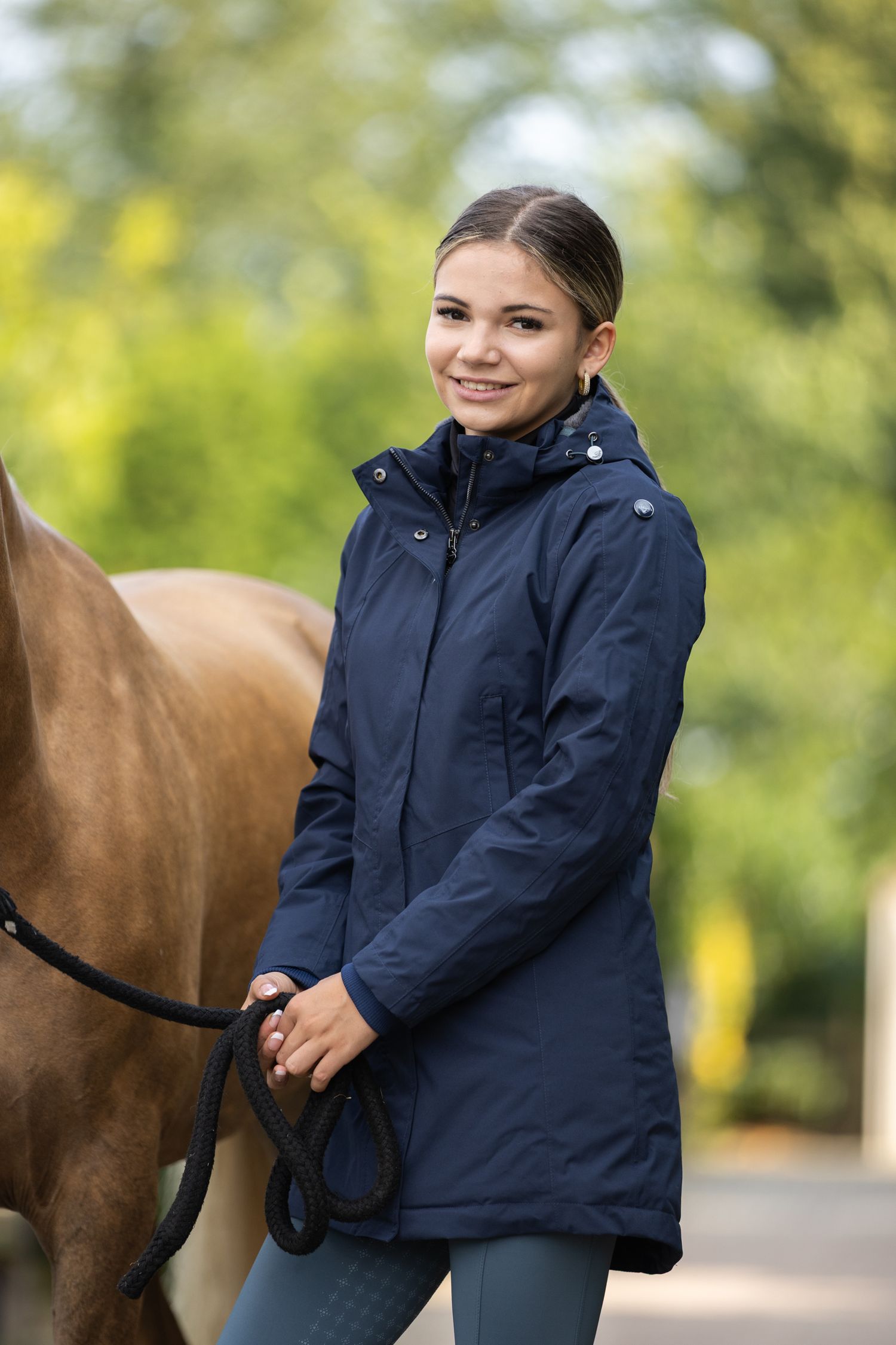 A rider in a navy jacket stands beside a brown horse outdoors.