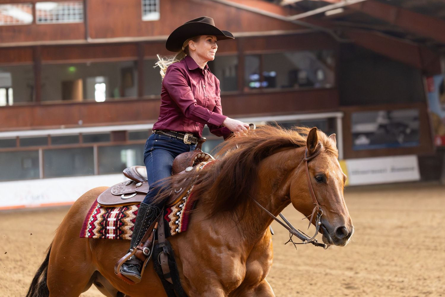 A rider in a burgundy shirt and cowboy hat guides a chestnut horse during a western riding competition in an indoor arena.