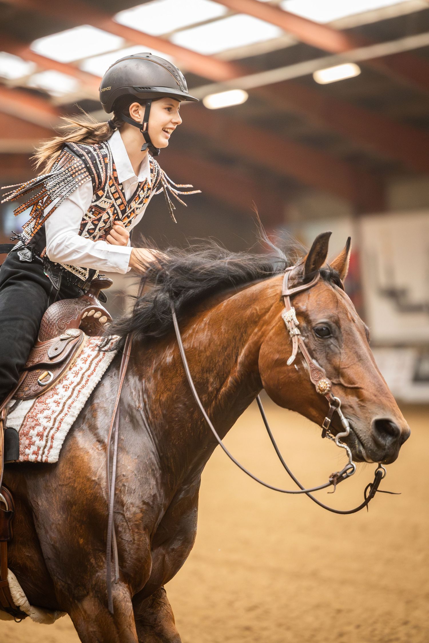 Smiling young female equestrian in helmet rides bay horse in indoor arena wearing decorative western vest.