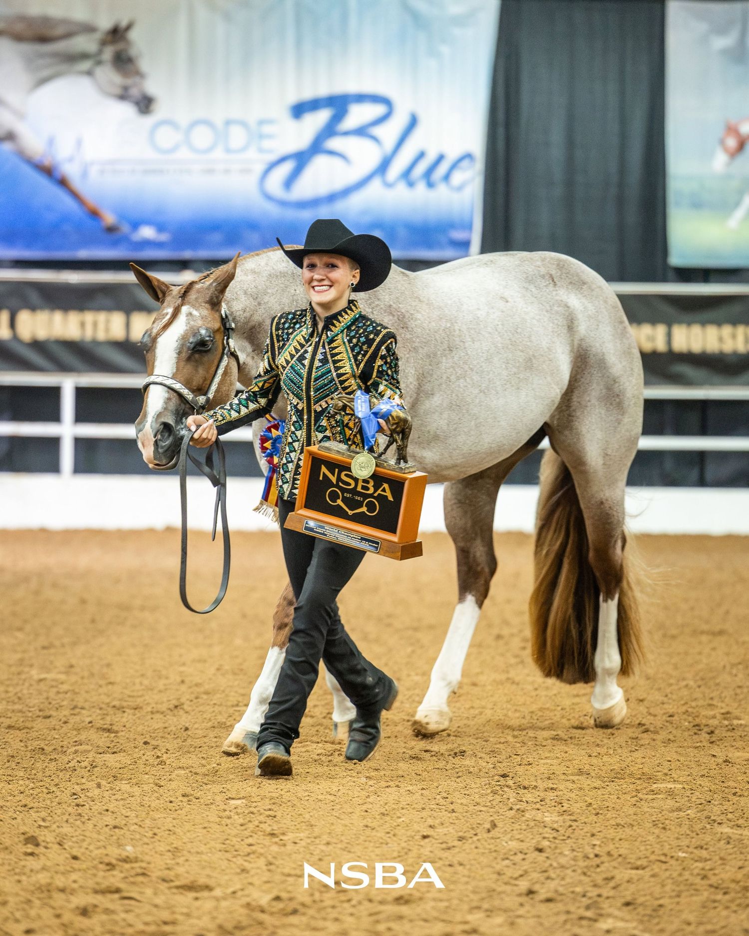 Young woman in western show attire smiles holding NSBA award trophy beside gray horse at the NSBA World Show