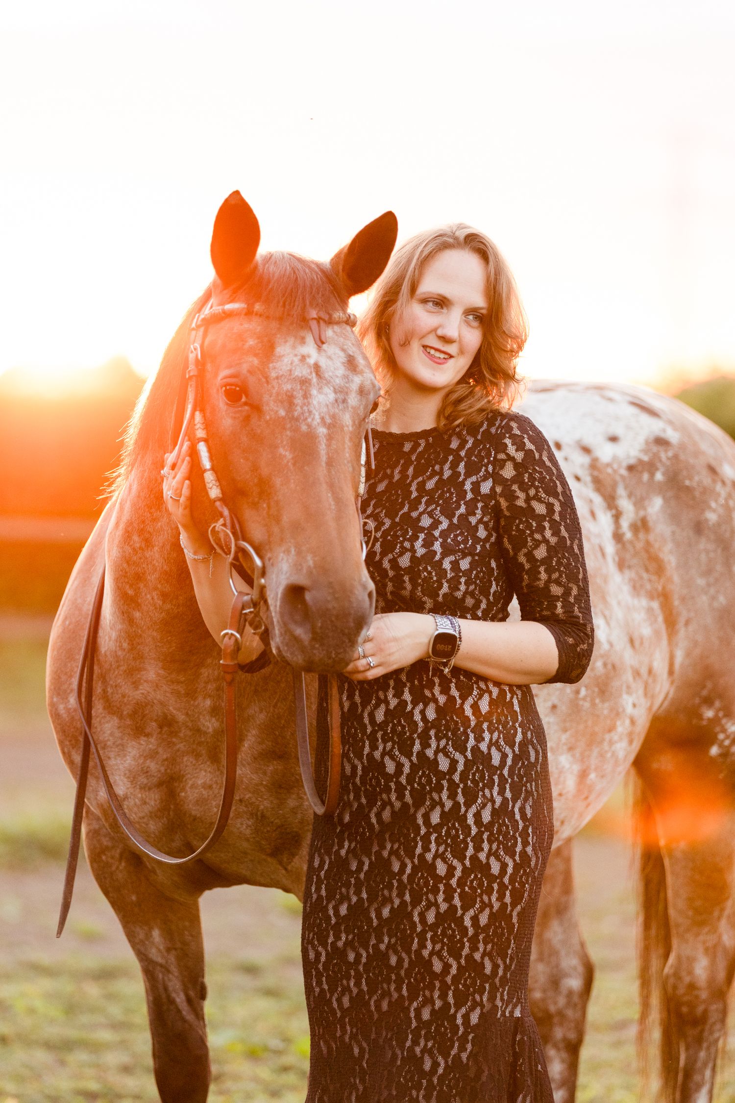 A spotted horse poses with someone in black lace against a warm sunset background.