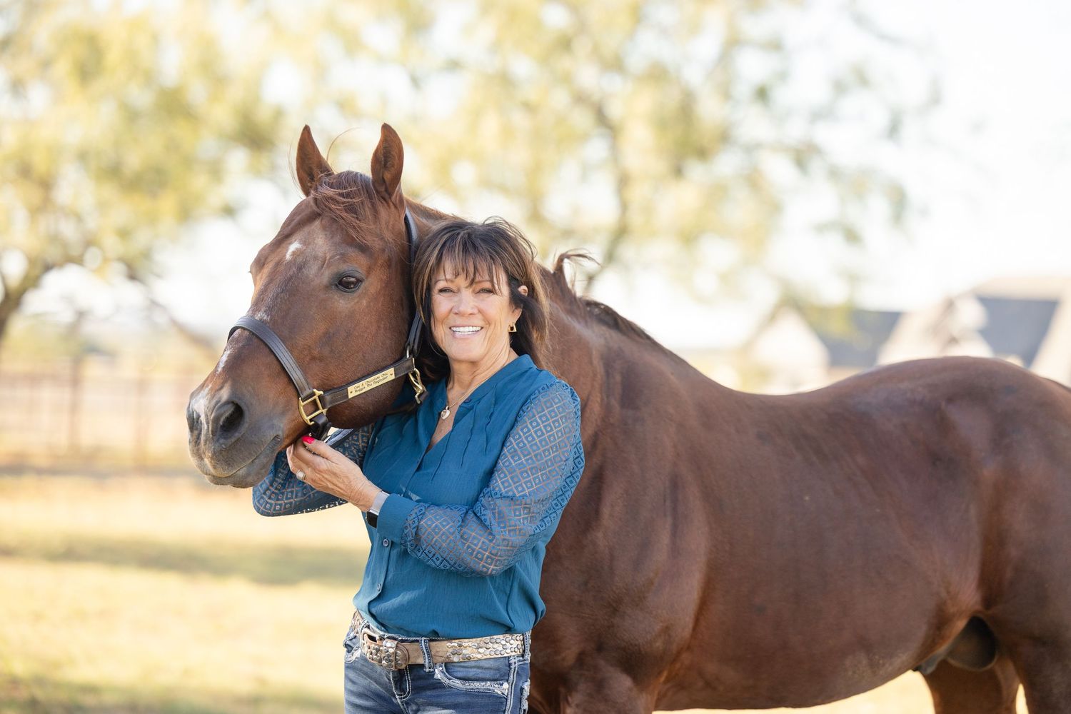 Person in denim shirt smiling while standing with brown horse in sunlit field.
